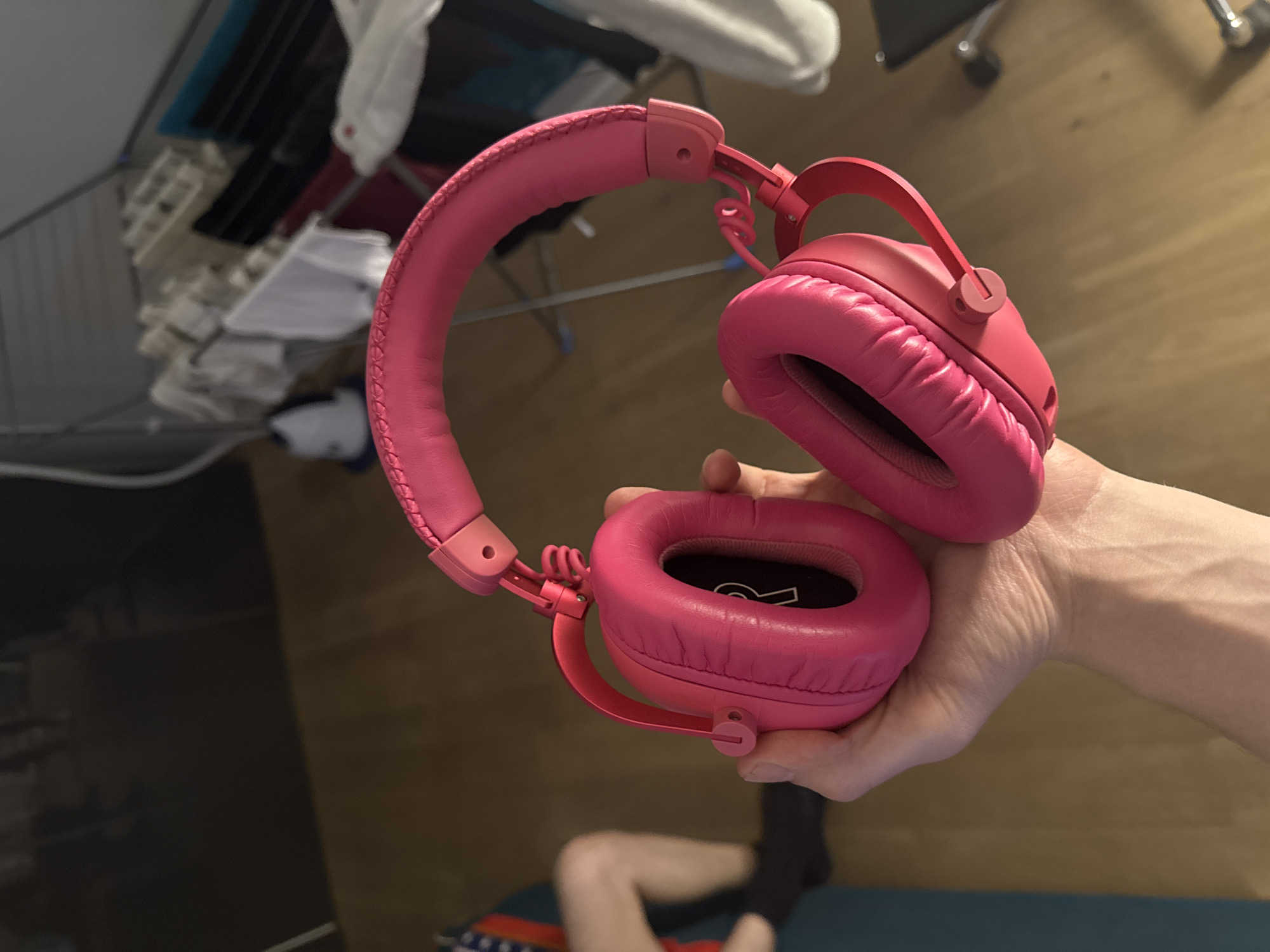 A person's hand holds a pair of bright pink over-ear headphones in an indoor setting. The headphones are the primary subject, with a padded headband and earcups. The background is blurred but shows what appears to be a room with wooden flooring, a drying rack with clothes, and furniture. A person's lower legs are visible in the left portion of the image.