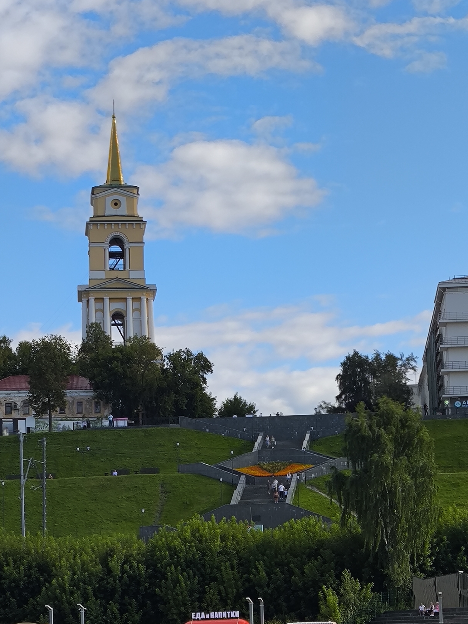 The image depicts a view of the city of Perm, Russia, focusing on the Church of the Kazan icon of the Mother of God bell tower, which is a prominent landmark. The bell tower, painted in light yellow with white accents and a golden spire, stands tall against a bright blue sky dotted with white clouds. In the foreground, a lush green hill slopes upwards, adorned with a winding staircase and vibrant flower arrangements. Several people are visible on the stairs and the grassy slopes, some walking and others resting. To the right of the bell tower, a modern white building with balconies is partially visible, indicating the urban environment. The overall scene suggests a pleasant day in a public space within the city.