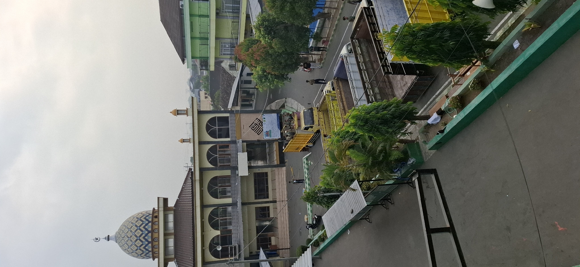 The image depicts an outdoor scene in the city of Wonorejo, Indonesia, featuring a mosque with a prominent blue and white checkered dome. The mosque building has a beige exterior with arched windows and a dark gray base. Adjacent to the mosque, a multi-story green building with balconies is visible. In the foreground and middle ground, several yellow and gray trucks are parked on a paved area. Some of these trucks are empty, while others appear to be carrying people or cargo. Several individuals are standing or walking around the trucks and the mosque. The environment includes trees, planters with greenery, and a walkway. The sky is overcast, indicating a cloudy day.