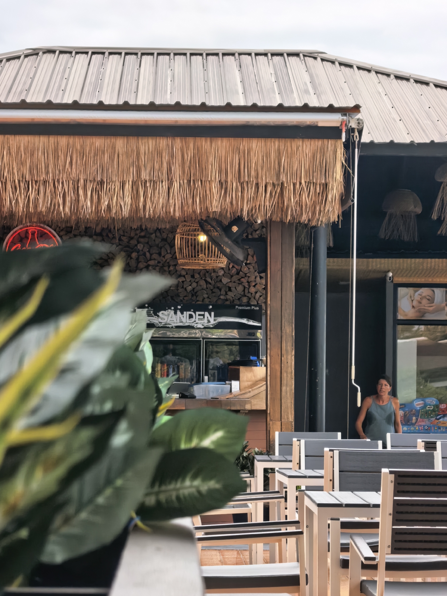 The image depicts an outdoor seating area in front of a building with a thatched roof overhang. A woman in a teal tank top stands near a doorway on the right side of the frame. Behind her, a glass display case shows various items. The building's facade features a stack of firewood and a neon sign. In the foreground, a large, out-of-focus green plant partially obscures the view of the seating area, which consists of multiple tables and chairs with grey and white accents. The scene appears to be located in Ko Samui District, Thailand.