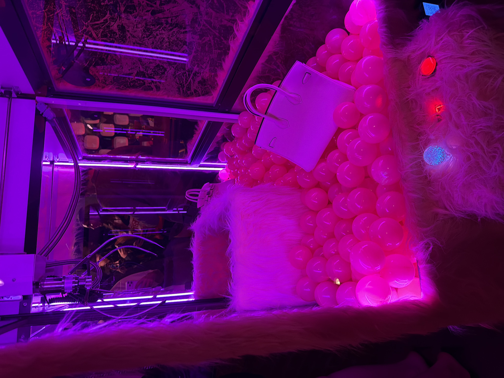 The image appears to be a display or installation, possibly in a shop window, featuring a white handbag propped against a mound of pink balloons. The scene is lit with strong pink and purple lighting. Fluffy white material is prominent, appearing as part of the display and possibly covering surfaces.
The background shows structural elements resembling the interior framework of a box or machine. There are reflective surfaces and geometric shapes, with pink LED lights illuminating the back interior.
Based on the presence of Russian text visible as a QR code tag on the upper right of the image, the location is Moscow, Russia.