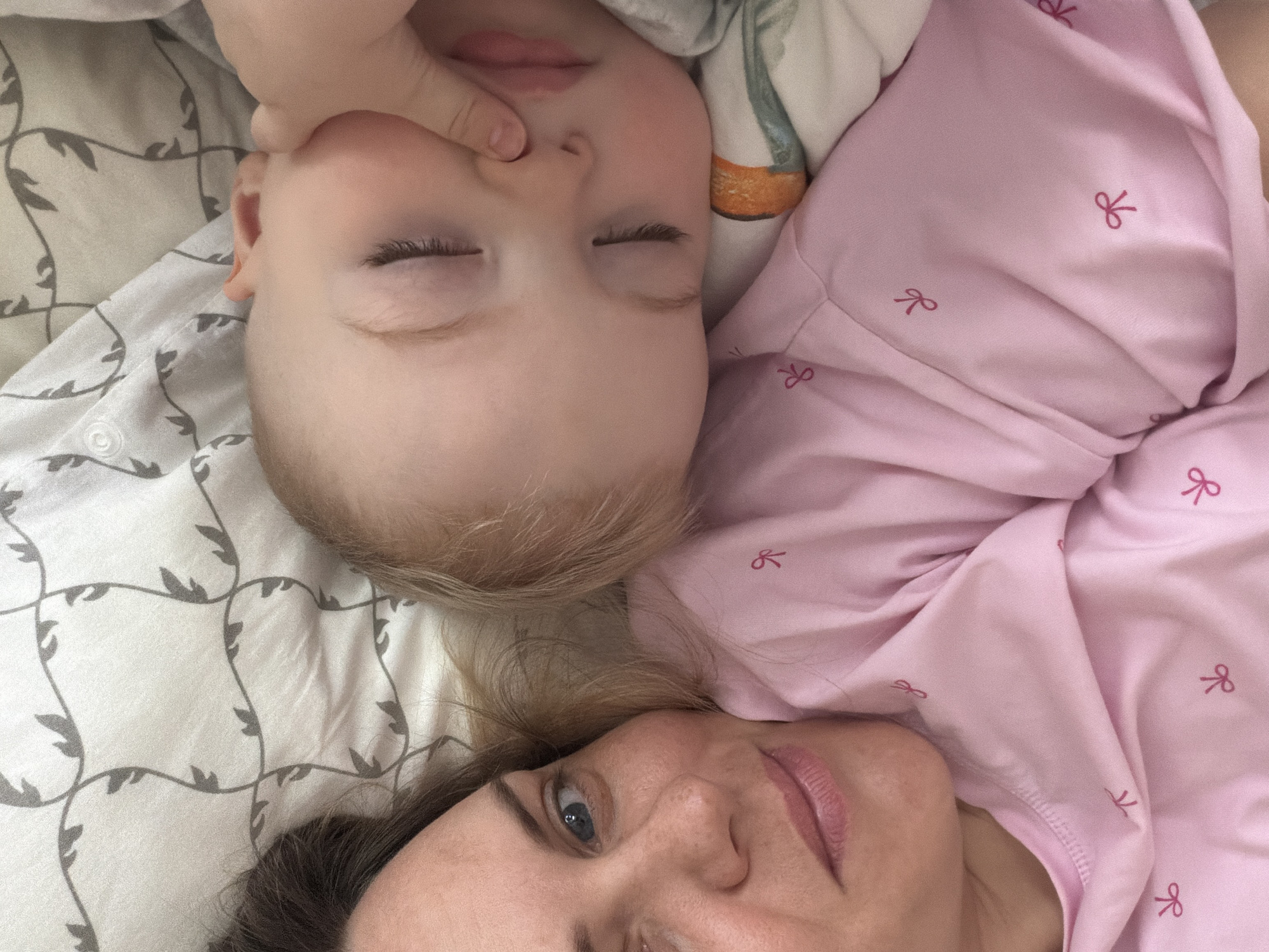 A woman and a young child are lying on a bed. The woman, positioned at the bottom of the frame, has her head tilted towards the camera, revealing part of her face. She has light skin, blondish-brown hair, and blue eyes. She is wearing a pink top adorned with small bow designs. The child, positioned above her, is lying on their back with closed eyes. A small hand is resting near the child's face. The bedding features a light-colored fabric with a subtle grey leaf pattern. The location of the image within the city of Votkinsk, Russia cannot be determined with the available information.