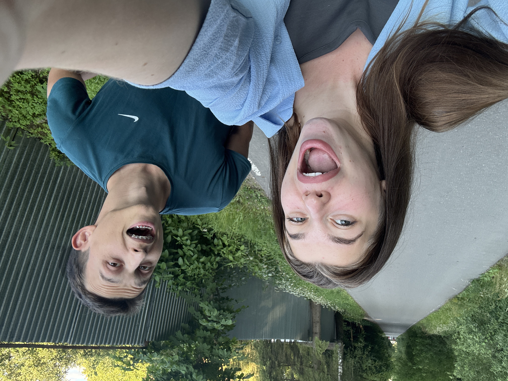 This is an upside-down, eye-level, selfie-style photograph featuring two young adults outdoors. The person on the left, a male with short brown hair and braces, is wearing a dark teal Nike t-shirt. He is looking directly at the camera with his mouth open in a surprised or playful expression. The person on the right, a female with long, dark brown hair, is wearing a grey t-shirt and a light blue garment draped over her shoulders. She also has her mouth wide open, facing the camera with a similar playful expression.
The background suggests an outdoor setting with lush green foliage and a grey, corrugated metal fence on the left. To the right, a paved path or sidewalk is visible, bordered by grass and wildflowers. The lighting indicates it is daytime, likely with bright sunlight filtering through the trees, creating dappled shadows. The overall mood of the image is lighthearted and jovial, with both individuals appearing to be sharing a moment of fun. The location context suggests this was taken in Shevlugino, Russia. No visible text is present in the image.