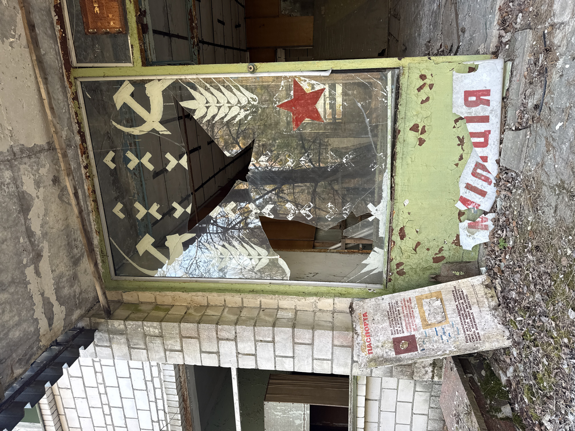 The image depicts an abandoned storefront in Prypiat, Ukraine, with a broken window displaying faded Soviet-era symbols: a hammer and sickle, a red star, and stylized wheat stalks. The surrounding structure shows signs of decay, including peeling paint, crumbling brickwork, and debris scattered around the entrance. A sign fragment with Cyrillic text, possibly related to passport application procedures ("ПАСПОРТА," indicating "passports"), lies on the ground, alongside other discarded paper debris. The overall scene suggests a post-abandonment environment, with no visible signs of human presence.