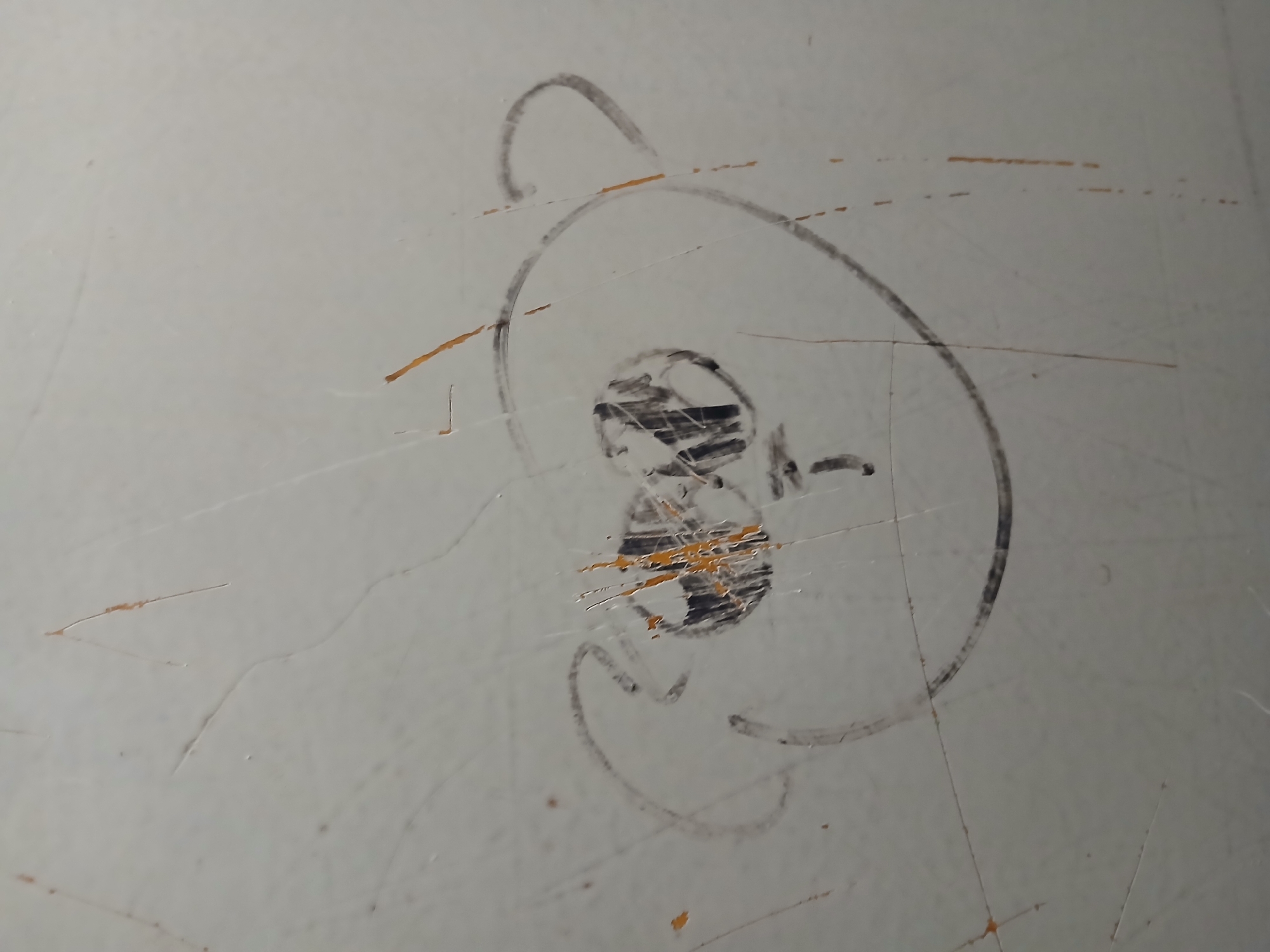 The image shows a rough, hand-drawn sketch of a face, possibly a cartoon character, on a scuffed, off-white surface. The face is outlined by a circular shape with two smaller circles for ears. Inside the large circle, two circular shapes represent eyes, with some black scribbling within them. A curved line below the eyes forms a mouth, and a small vertical line below the mouth could be a nose. The drawing is done in black marker or crayon. The surface itself is marred by numerous scratches and some brown discoloration, suggesting it is a wall or a similar textured surface. The overall impression is of graffiti or children's drawing on a damaged surface. The location in the city of Ukrainka, Ukraine, is not evident from the image itself.