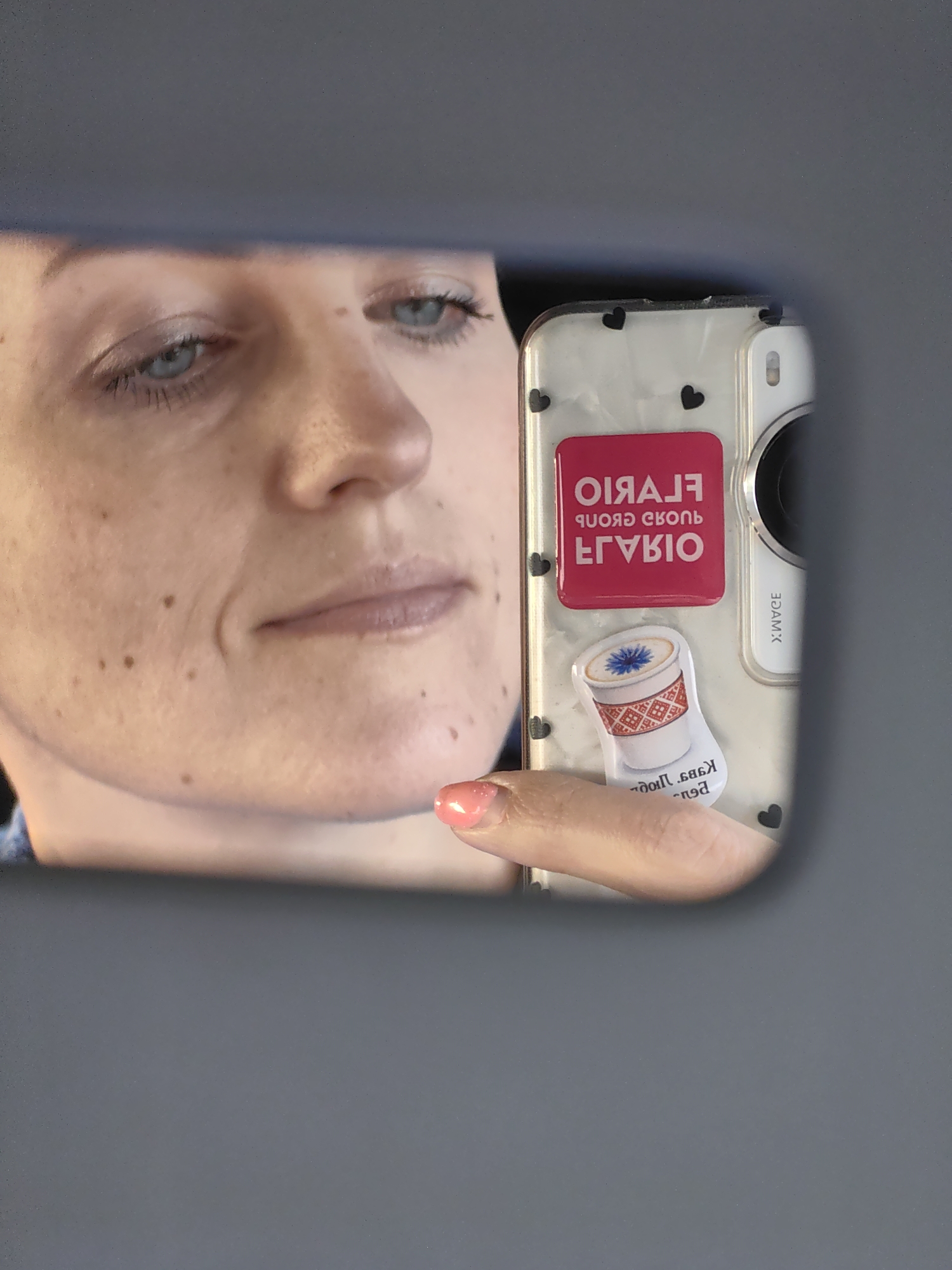 The image is a reflection in a car's rearview mirror, showing a person's face and a smartphone. The person has light-colored eyes, fair skin with freckles and moles, and is wearing light makeup. The smartphone is in a clear case with black heart designs. It has two stickers on it: one is a pink square with white text that appears to be a logo, and the other is a sticker of a coffee cup. The person's hand, with a pink nail polish, is visible holding the phone. The background is a blurred gray surface, possibly the interior of a car. The reflection shows the left side of the person's face in the foreground, with the right side of the face and the phone reflected in the mirror. The image was taken on March 13, 2024, at 11:30 AM in Fanipal, Belarus.