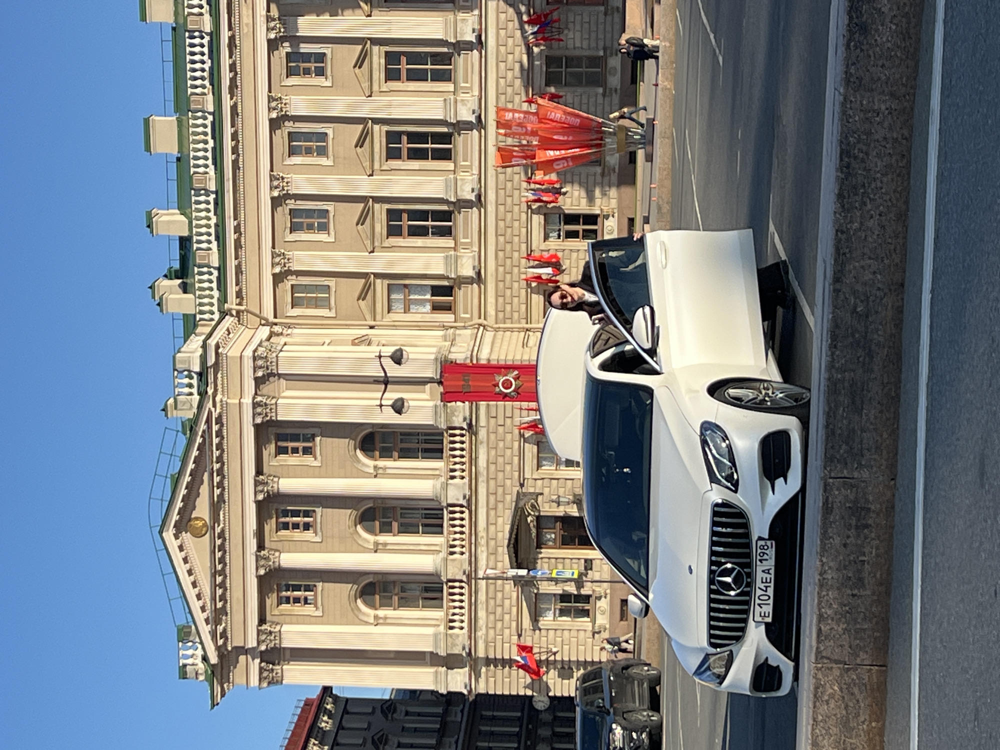 The image captures a street scene in Saint Petersburg, Russia. A white Mercedes-Benz is parked on the side of the road, with its driver's side door open, revealing a person in the driver's seat. The vehicle has a license plate that reads "E 104 EA 198." Behind the car stands a multi-story building with classical architecture, adorned with columns, ornate window frames, and decorative elements. Red flags and banners are displayed on the building's facade. People are seen on the sidewalk in the background. The sky is clear and blue.