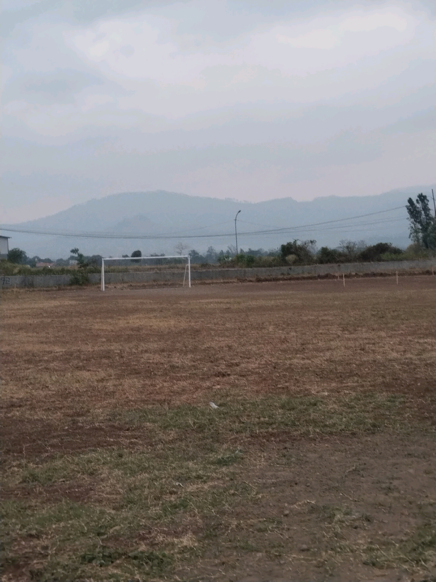 This is a landscape photograph of a dry, unkempt soccer field in Jembayat, Indonesia. The field is covered in sparse, brown grass and dirt, with some patches of greener growth. A white soccer goal is visible in the middle ground, positioned towards the left side of the frame. In the background, there is a low concrete wall, beyond which are some trees and buildings. A hazy, mountainous landscape forms the horizon, under a pale, overcast sky. The overall impression is one of neglect and a lack of recent activity.