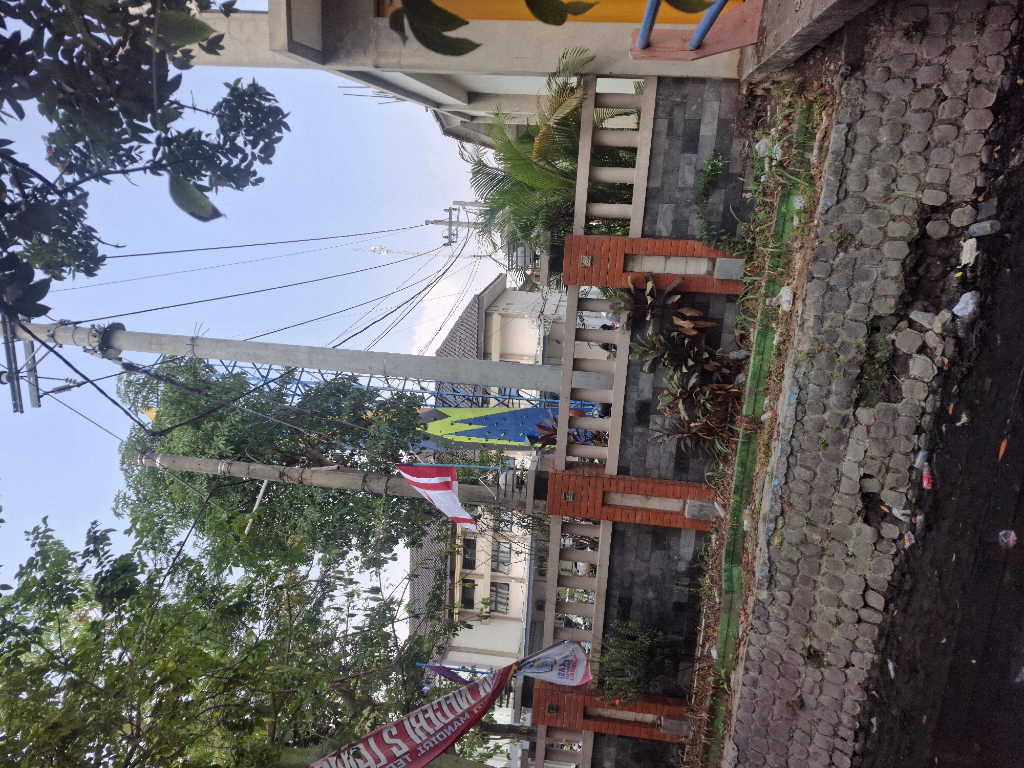 The image depicts a street scene in Jetis Indah, Indonesia. A narrow canal runs along the bottom of the frame, its water dark and littered with plastic bottles and debris. The canal walls are constructed of rough, gray stones. Above the canal, a fence made of gray stone and red brick pillars separates it from a walkway. Beyond the fence, several buildings are visible, including a light-colored structure with dark-framed windows and a tiled roof.
To the left, a red banner with white lettering is draped over a tree. A white and red flag, possibly the Indonesian flag, flies from a utility pole in the center of the image. A colorful climbing wall, with blue and yellow sections, stands behind the flag. Trees with green foliage are scattered throughout the scene, some partially obscuring the sky, which is a pale blue with scattered clouds. Electrical wires crisscross the sky, supported by utility poles. The overall impression is one of an urban environment with signs of neglect and litter.