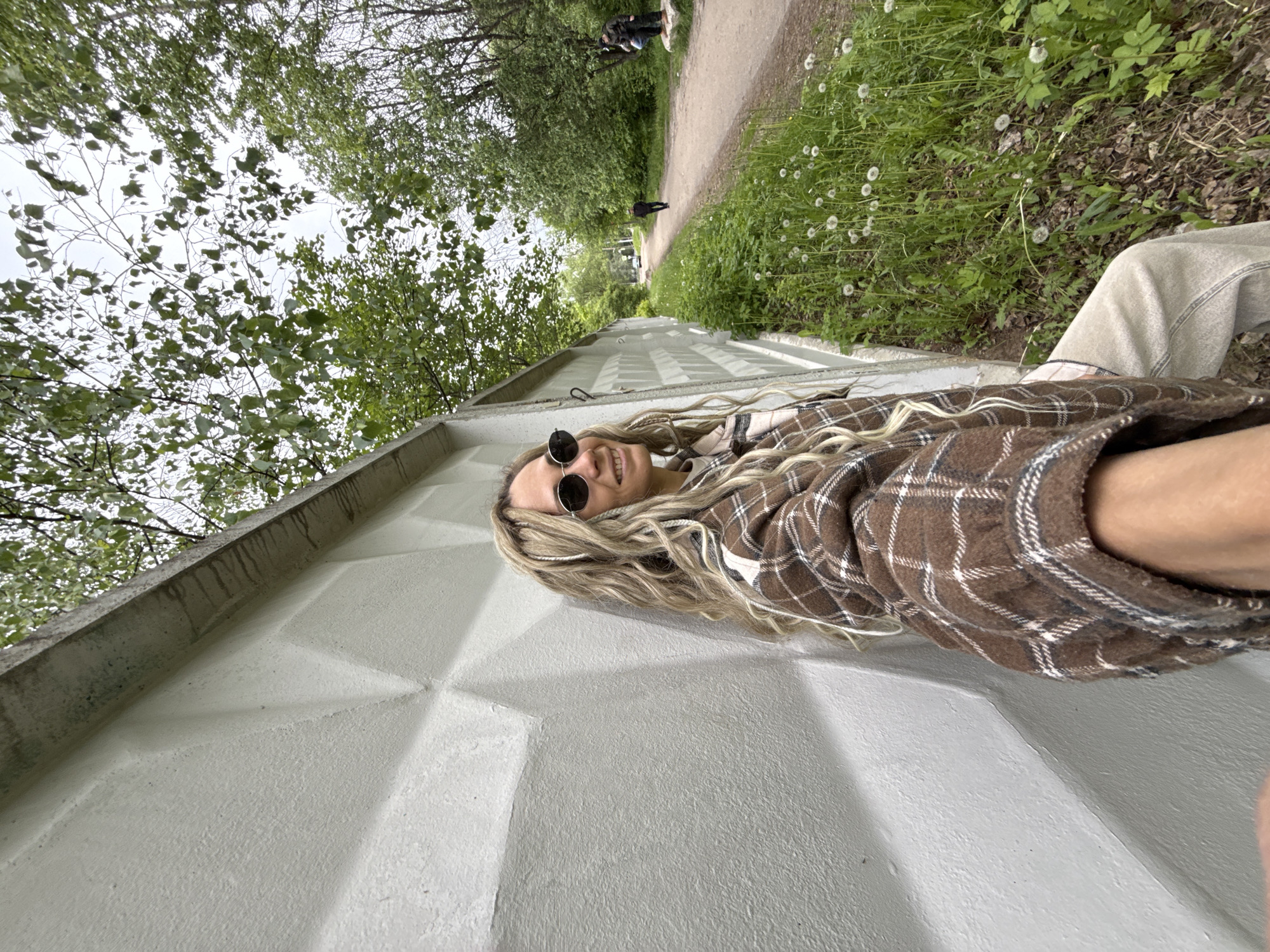 A photo shows a person lying on a gray concrete wall in Rusanovo, Russia. The person has long blonde hair, wears sunglasses and a brown plaid shirt. A pathway with trees and grass can be seen in the background. There are other people further down the path.