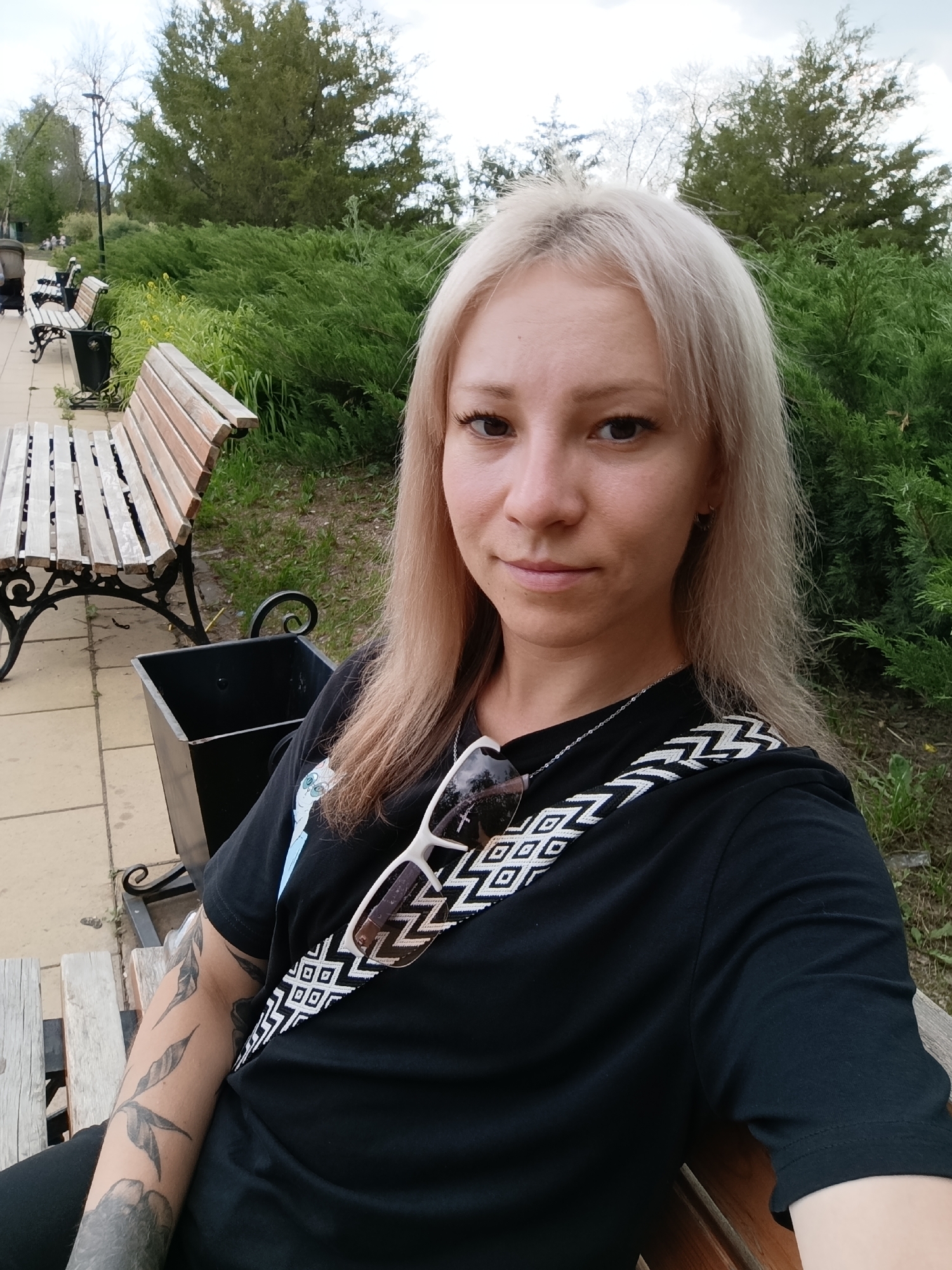 A blonde woman with tattoos on her left arm is sitting on a wooden bench in a park, taking a selfie. She is wearing a black t-shirt and sunglasses are resting on her shirt. A black trash can is visible next to the bench. The park has trees and bushes, and the ground is paved with light-colored tiles. In the background, there are more benches and a stroller. The image appears to have been taken in Novokuybyshevsk, Russia, given the context.