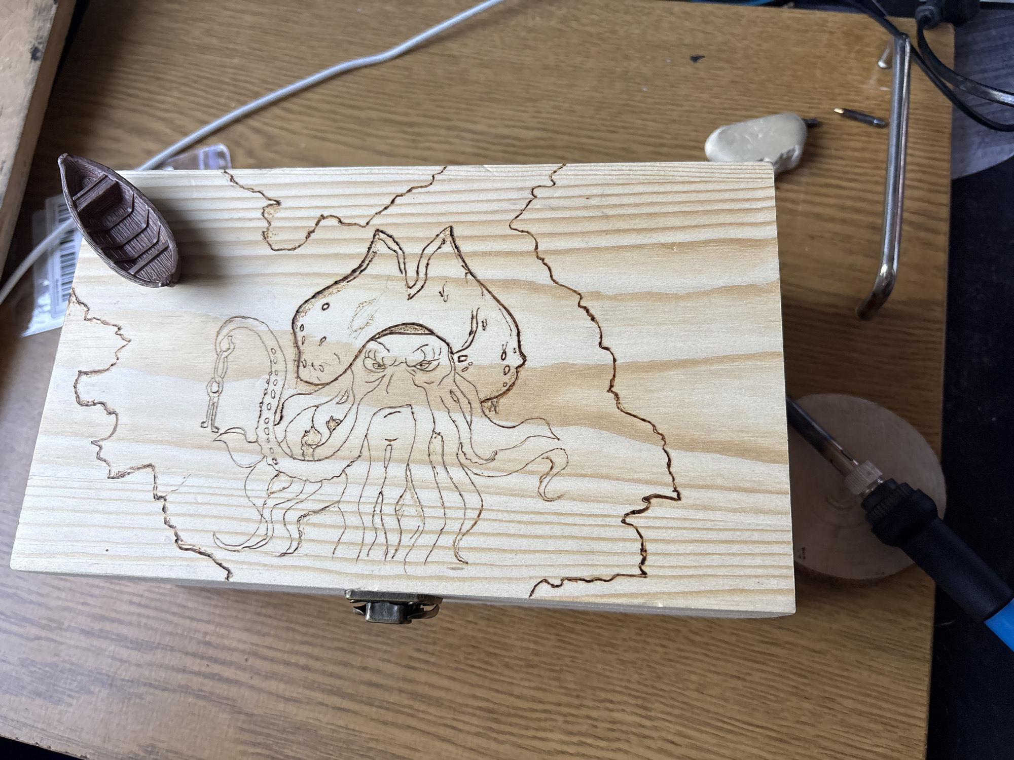 The image shows a light-colored wooden box with a burned-in design of Davy Jones from Pirates of the Caribbean within an outline resembling a map. A small, dark brown rowboat sits on the upper left corner of the box. The box is positioned on a light brown wooden surface. Various objects surround the box: a white cable, a small package, a white putty-like substance, a silver tool, a wooden disc, and a blue and black wood-burning tool. The background includes glimpses of dark and light brown surfaces. The location is not specified in the image. There is no information in the image to indicate its location in Sofia, Bulgaria.