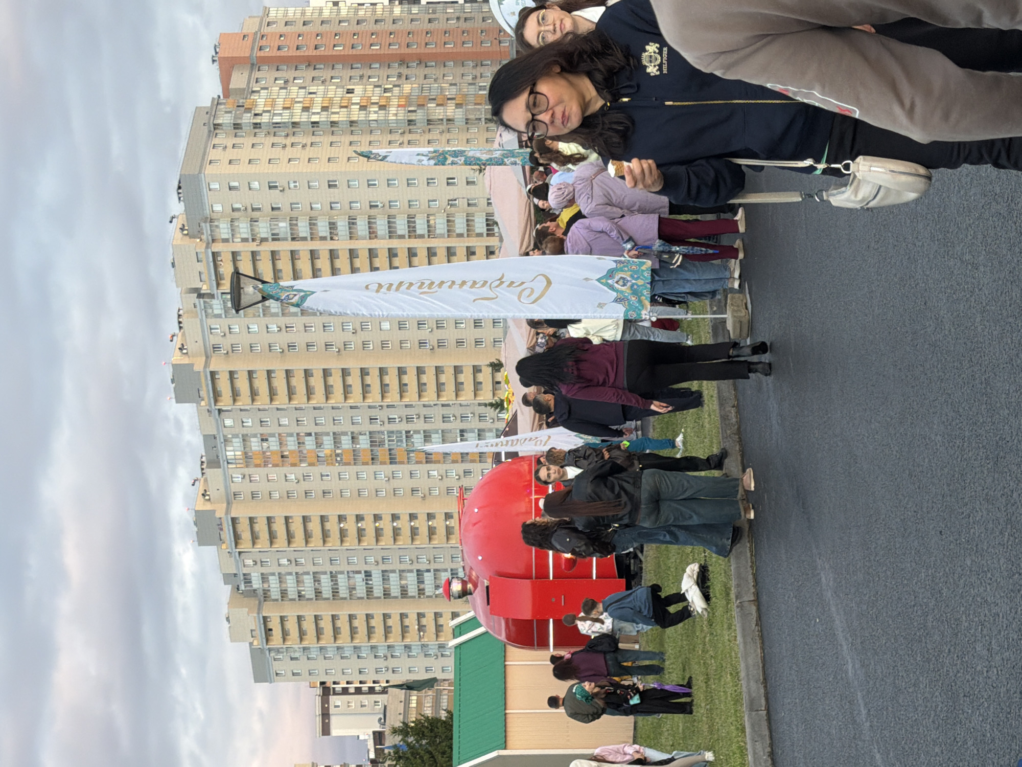 The image shows an outdoor scene with a crowd of people gathered in front of a bright red, rounded structure that resembles a stylized fire truck. In the background, there are tall, multi-story apartment buildings with many windows. The sky is cloudy. Several white flags with decorative patterns and text are visible among the crowd. People are standing and walking around, some are looking at the fire truck structure, while others are engaged in conversation. One woman in the foreground is holding an ice cream cone. The ground is paved, and there is a strip of grass with a curb separating it from the pavement. The overall impression is that of a public gathering or festival.