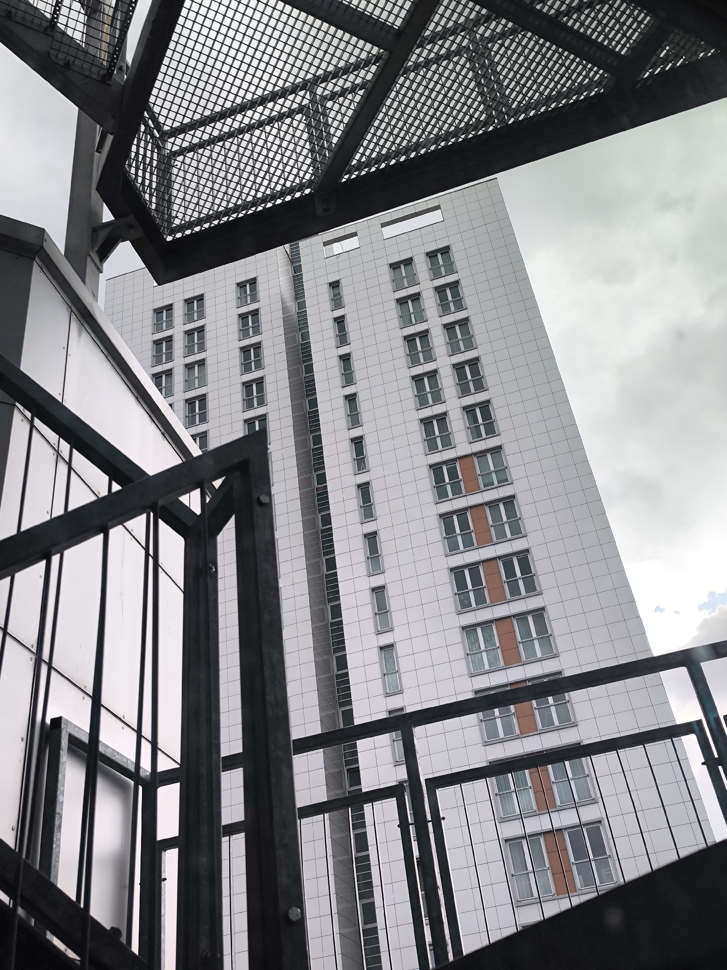 The image captures a low-angle view in Riga, Latvia, presenting a modern architectural setting. The foreground is dominated by industrial-style metal stairs and railings, constructed from dark metal. Above the stairs, a grating structure filters the light. The main subject is a tall, slender skyscraper with a grid-like facade of white panels interspersed with rows of windows. Some window sections are accented with orange panels. The sky is overcast, creating a diffused light across the scene. The image suggests an urban environment.