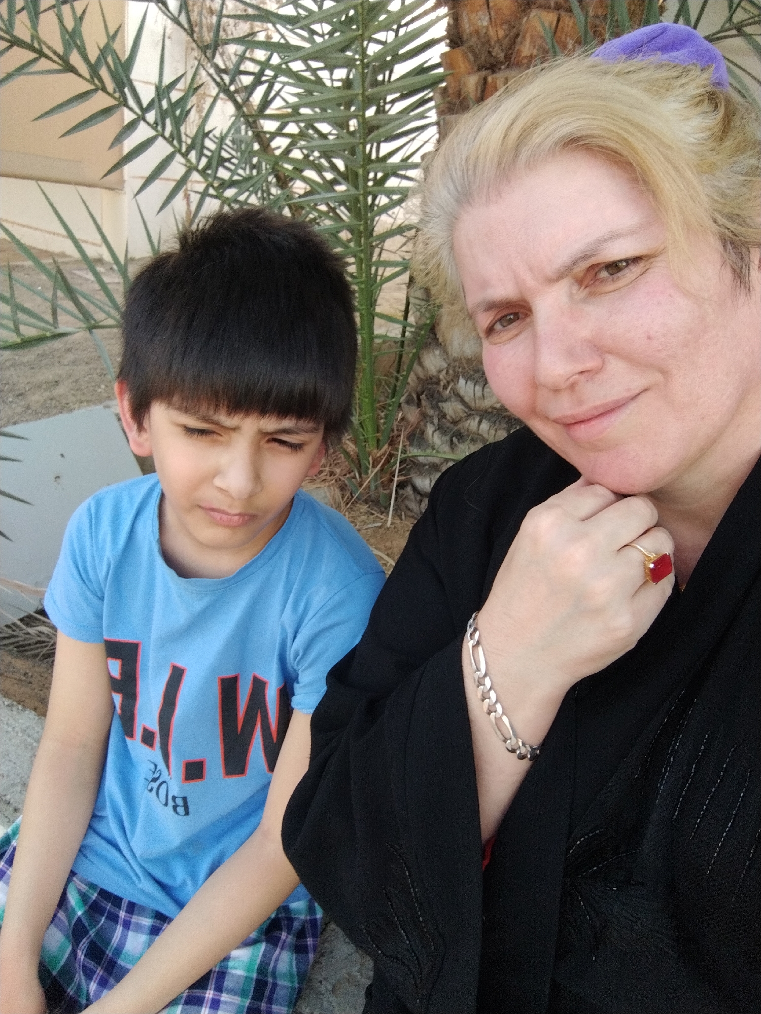 A woman and a young boy are posing for a photo outdoors. The woman is on the right, wearing a black garment and a purple hair covering. She has blonde hair and fair skin, and she is resting her chin on her hand, which has a silver bracelet and a ring with a red stone. The boy is on the left, wearing a blue t-shirt and plaid shorts. He has dark hair and appears to be frowning. Behind them, there's a palm tree and some green foliage, suggesting a natural or landscaped setting. The location is Al Mas'oudi, United Arab Emirates.