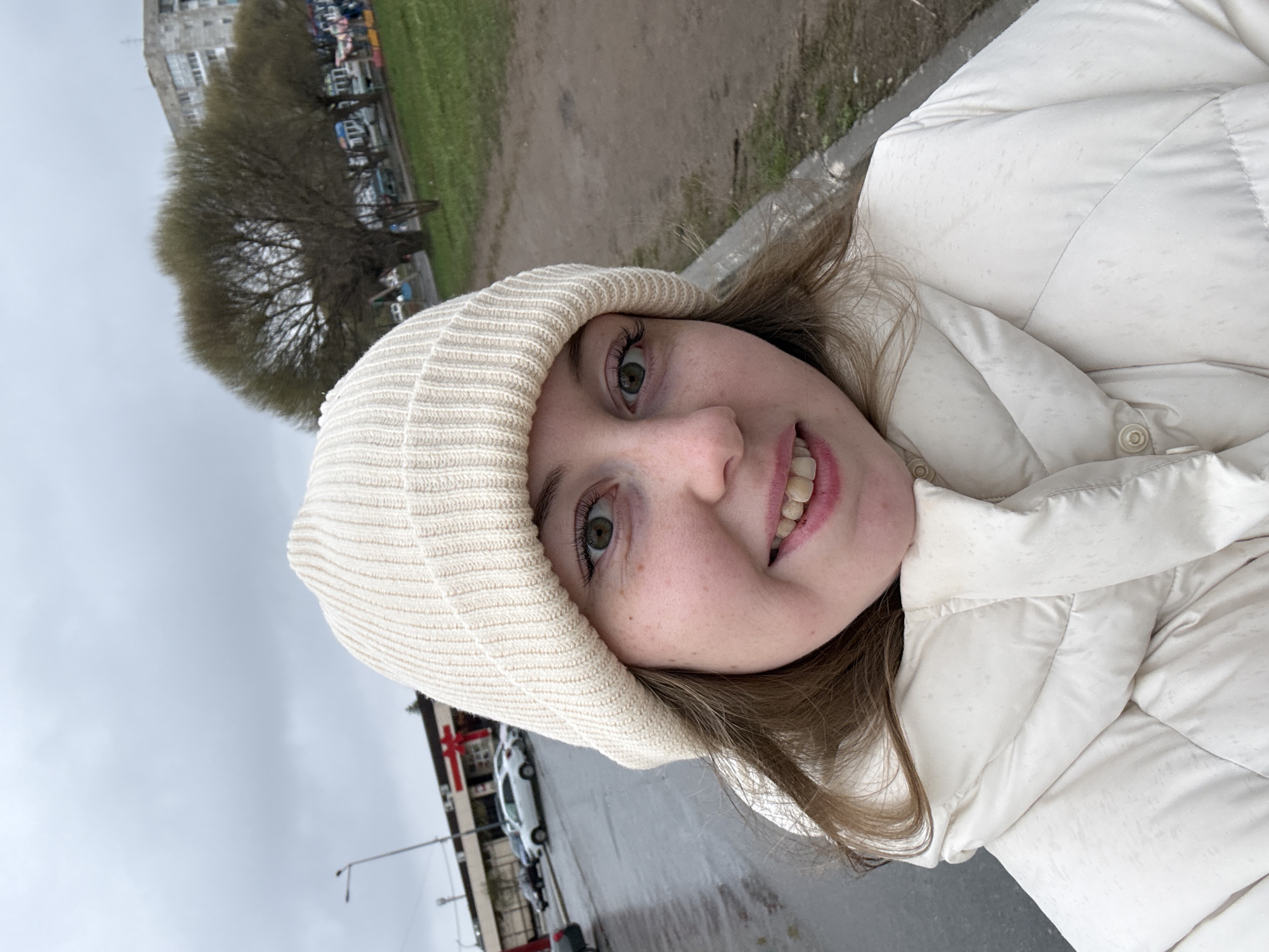 A selfie captures a young woman wearing a cream-colored knit hat and a matching puffer jacket. She is smiling and looking directly at the camera. The background reveals an urban environment. There is a tree and part of a building to the left. In the distance, cars are parked in front of a building. The sky is overcast. The location is Unknown, Unknown.