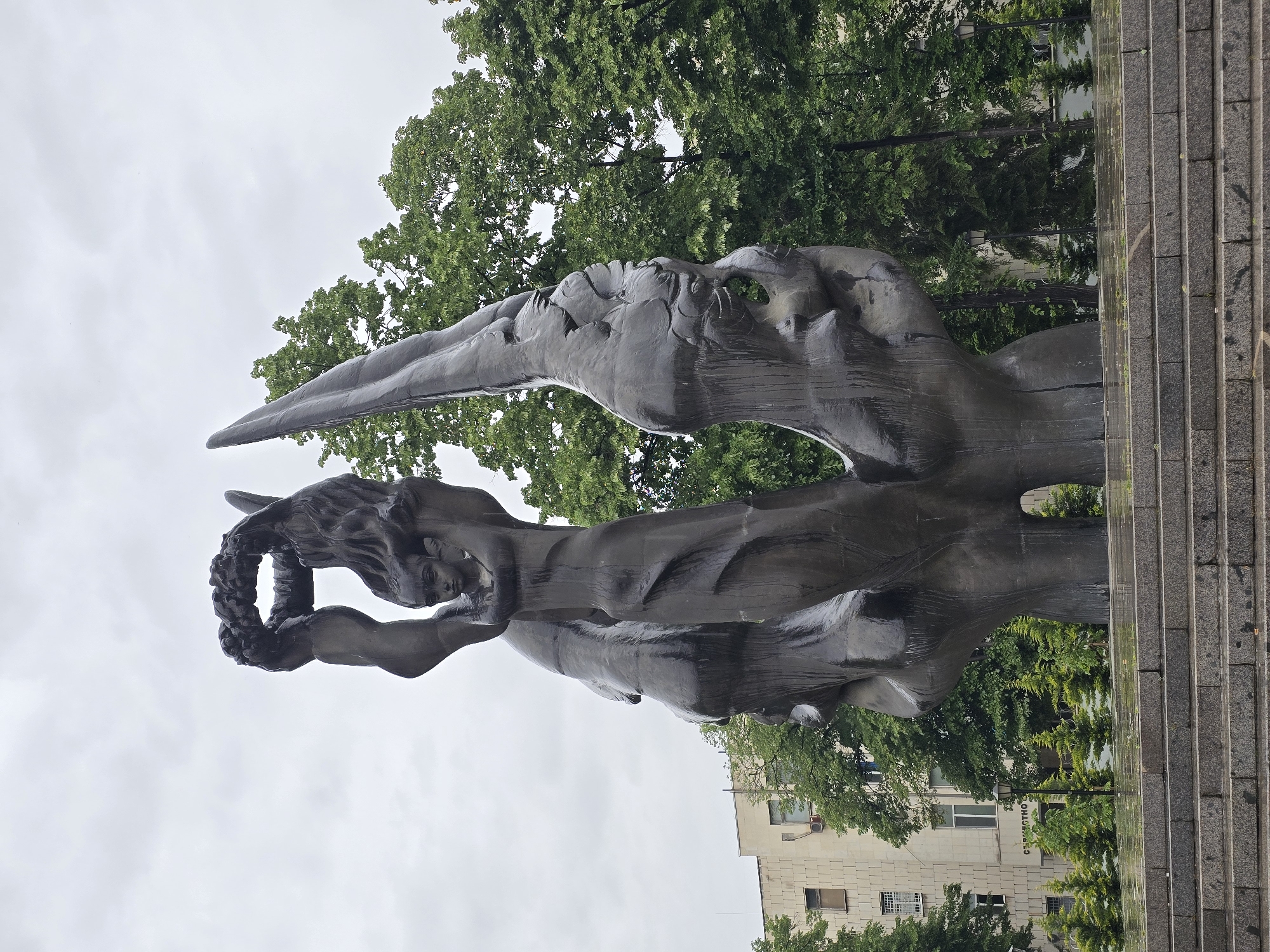 A bronze sculpture stands prominently in an outdoor urban setting of Unknown, Unknown. The sculpture depicts a winged figure raising a wreath. Green trees surround the sculpture, and a building is visible in the background. Stairs made of gray stone are on the right side of the sculpture. The sky is overcast.