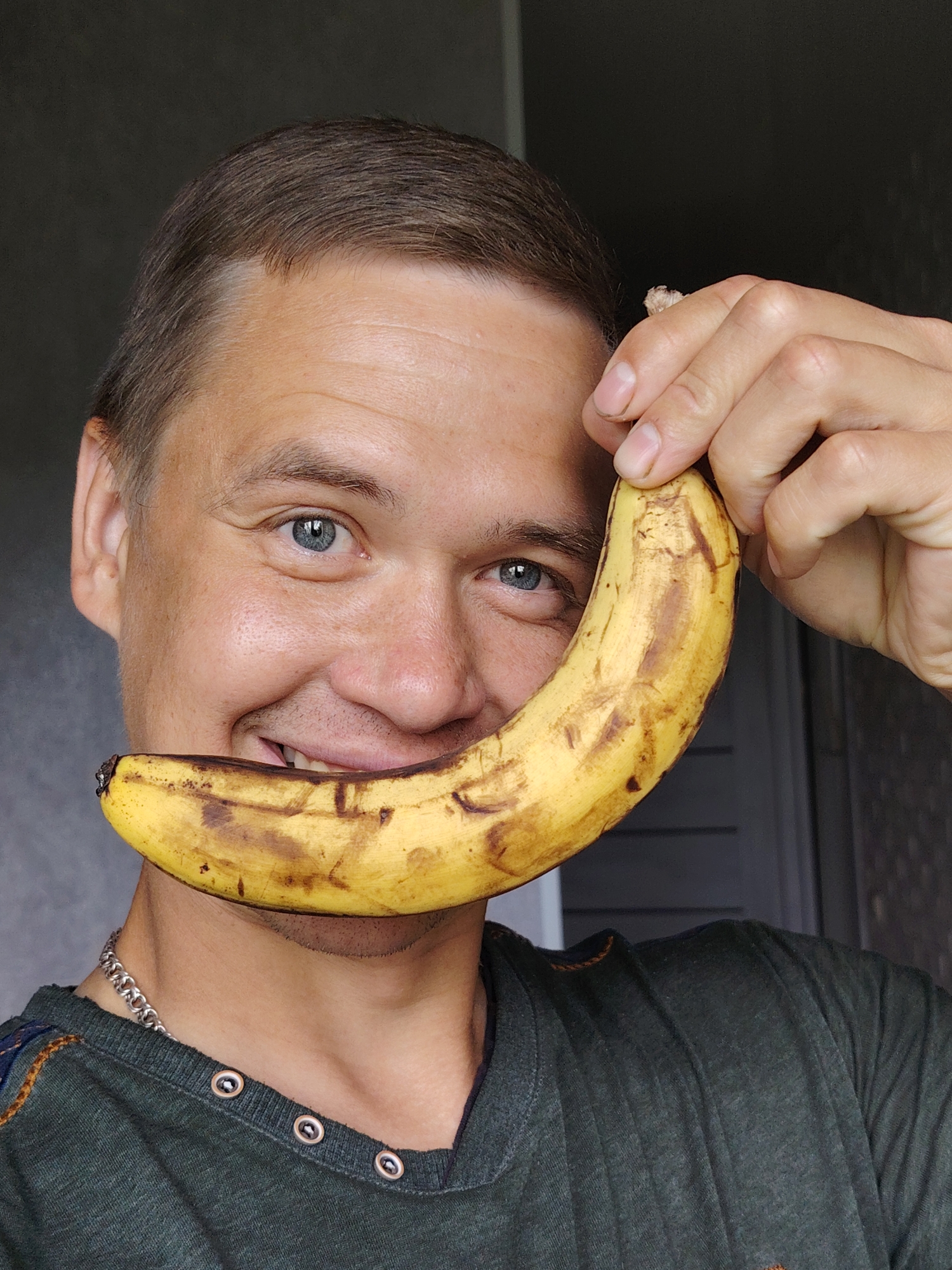 A close-up, head-and-shoulders shot shows a Caucasian man with fair skin, short brown hair, and blue eyes. He is smiling and holding a partially peeled, ripe yellow banana with brown spots up to his mouth, obscuring his lips. He is wearing a dark green t-shirt with a buttoned neckline and a silver chain necklace. The background is out of focus and appears to be an indoor environment with a dark wall and a door or window. The man is positioned in the center of the frame. The image was taken in the city of Chaikovsky, Russia.