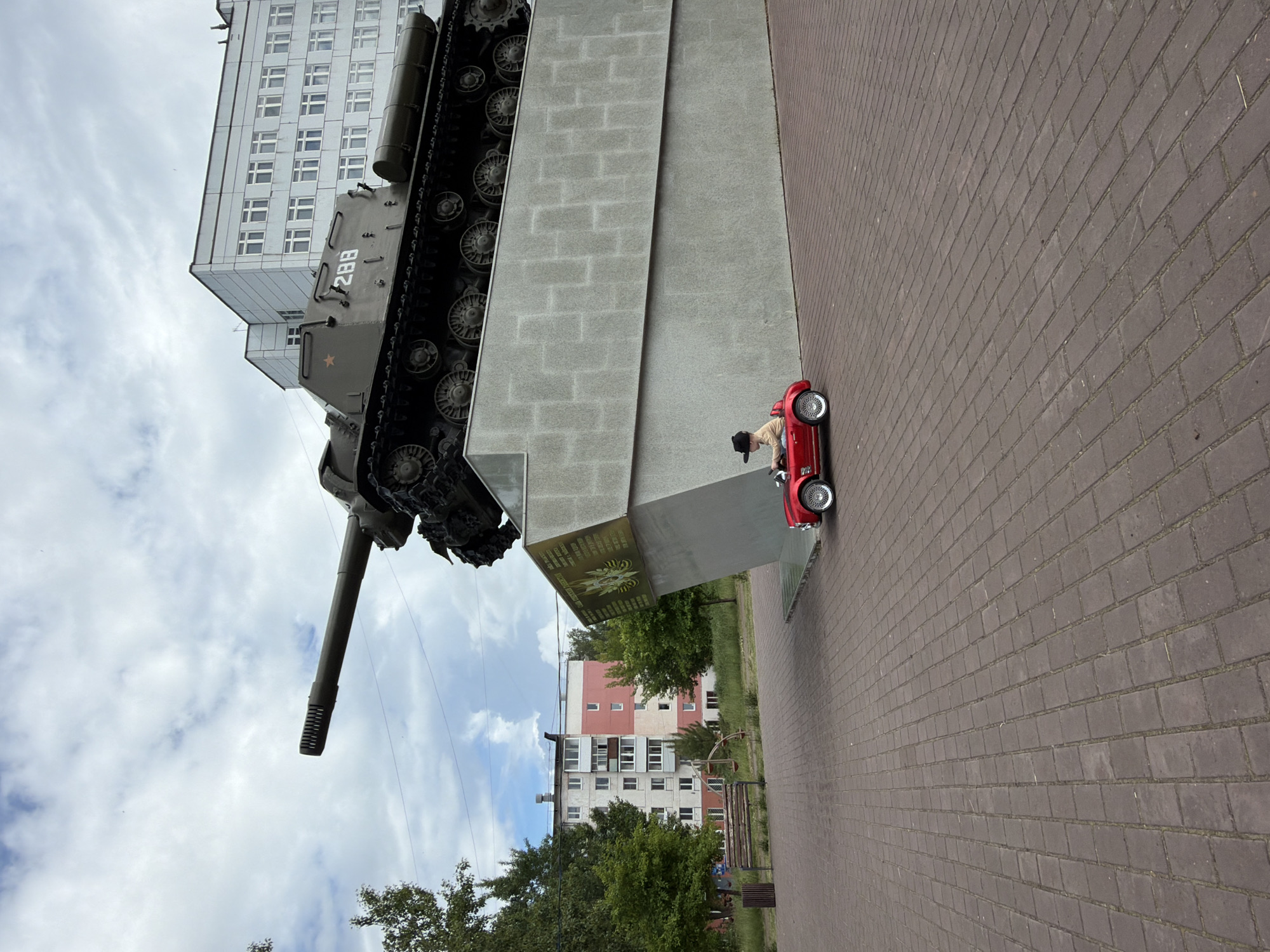 In Chaikovsky, Russia, a child sits in a toy red car on a paved area in front of a monument. The monument features a large tank mounted on a concrete pedestal. Behind the monument, a tall, multi-story building with a grid of windows stands under a cloudy sky. To the left of the monument, there are trees and apartment buildings. The foreground is a brick-paved plaza.