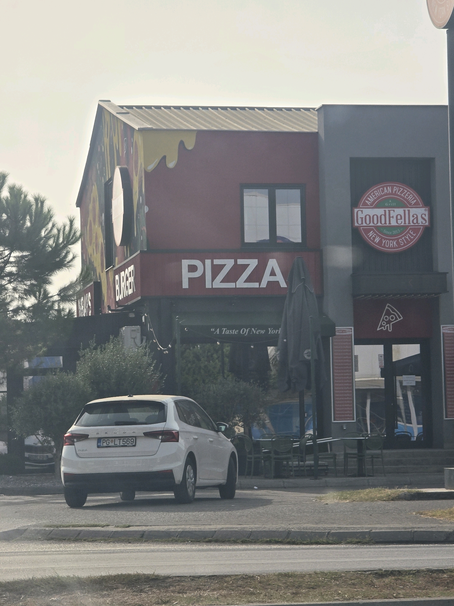 The image shows a white Skoda Fabia car parked on the side of a road in front of a commercial building. The building appears to be a restaurant with a large "PIZZA" sign and a circular "GoodFellas American Pizzeria New York Style" logo. The car has a license plate that reads "PG LT569". The environment is urban, with trees and other buildings visible in the background. The sky is overcast. It is likely the car is parked on a street in Podgorica, Montenegro, given the "PG" prefix on the license plate which is commonly used for Podgorica.