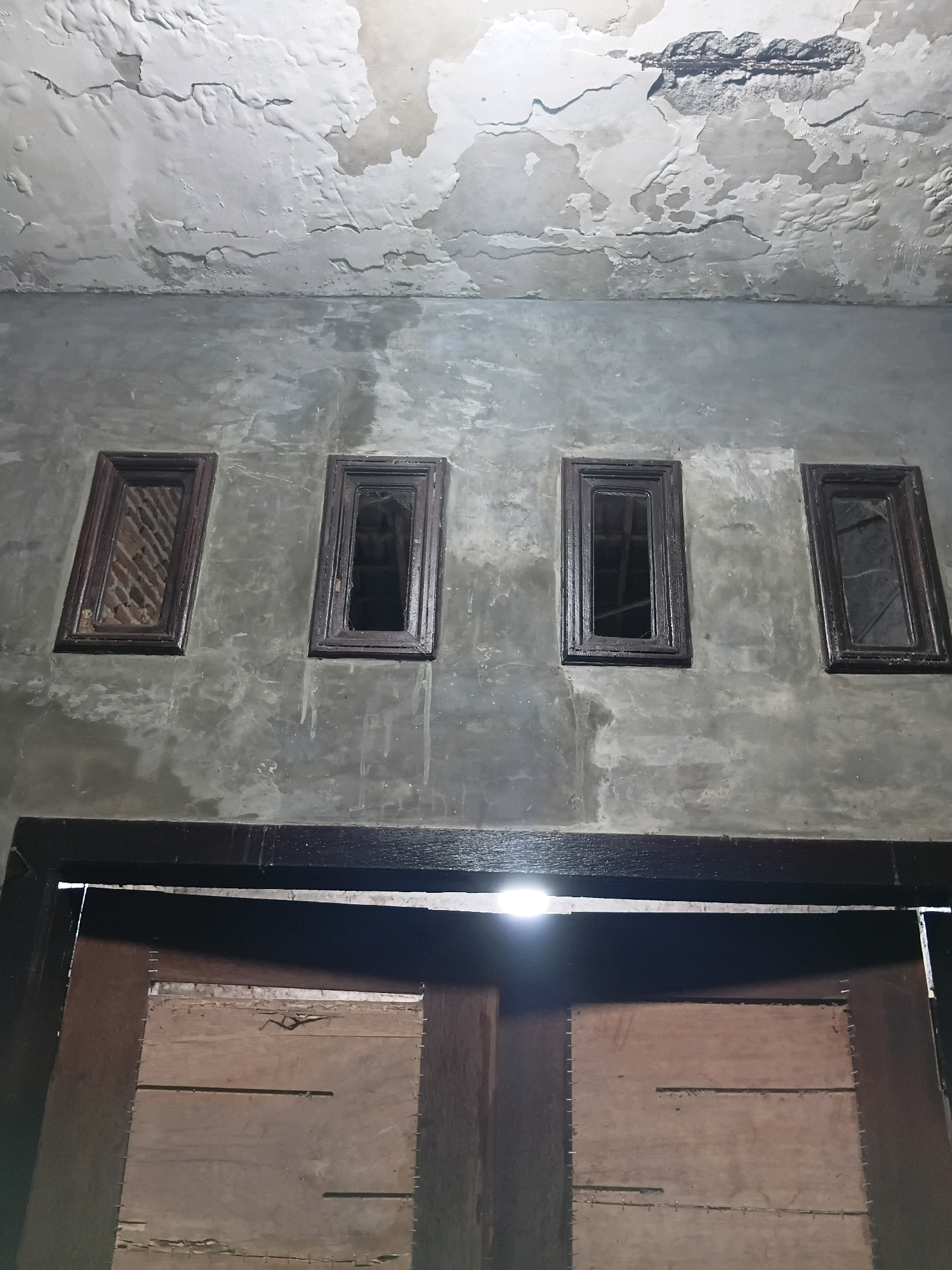 The image depicts a dark, weathered interior scene, likely in Cikal, Indonesia, characterized by signs of disrepair. The ceiling exhibits extensive damage, with peeling paint and exposed concrete revealing rebar. Below the damaged ceiling, a concrete wall features four small, dark-framed windows. The leftmost window shows a glimpse of brickwork, while the others are dark or reflect indistinctly. A wide, dark wooden beam runs horizontally across the image, partially obscuring a doorway. The doorway itself is made of wooden planks, with visible nails and some fraying material. A bright light source, possibly a lamp, illuminates a portion of the doorway from above. The overall impression is one of an old, neglected space.