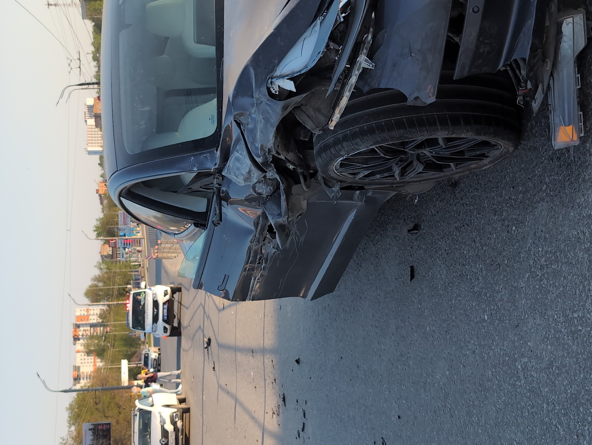 The image depicts the aftermath of a car accident on a city street. A dark-colored sedan has sustained significant damage to its front end, particularly on the driver's side. The front bumper is torn, the headlight is damaged, and the fender is crumpled. The front left tire is visible and appears intact.
In the background, multiple vehicles are present, including a white emergency vehicle, potentially a police car, with visible blue flashing lights. Several individuals are standing near the emergency vehicle, possibly observing the scene or involved in the accident response. Buildings are visible in the background, suggesting an urban setting. The license plate of one of the cars in the background is visible, but cannot be read.
The setting is identified as Izhevsk, Russia, based on the license plates in the background and the architecture of the buildings. The time of day appears to be daytime. The road surface is asphalt.