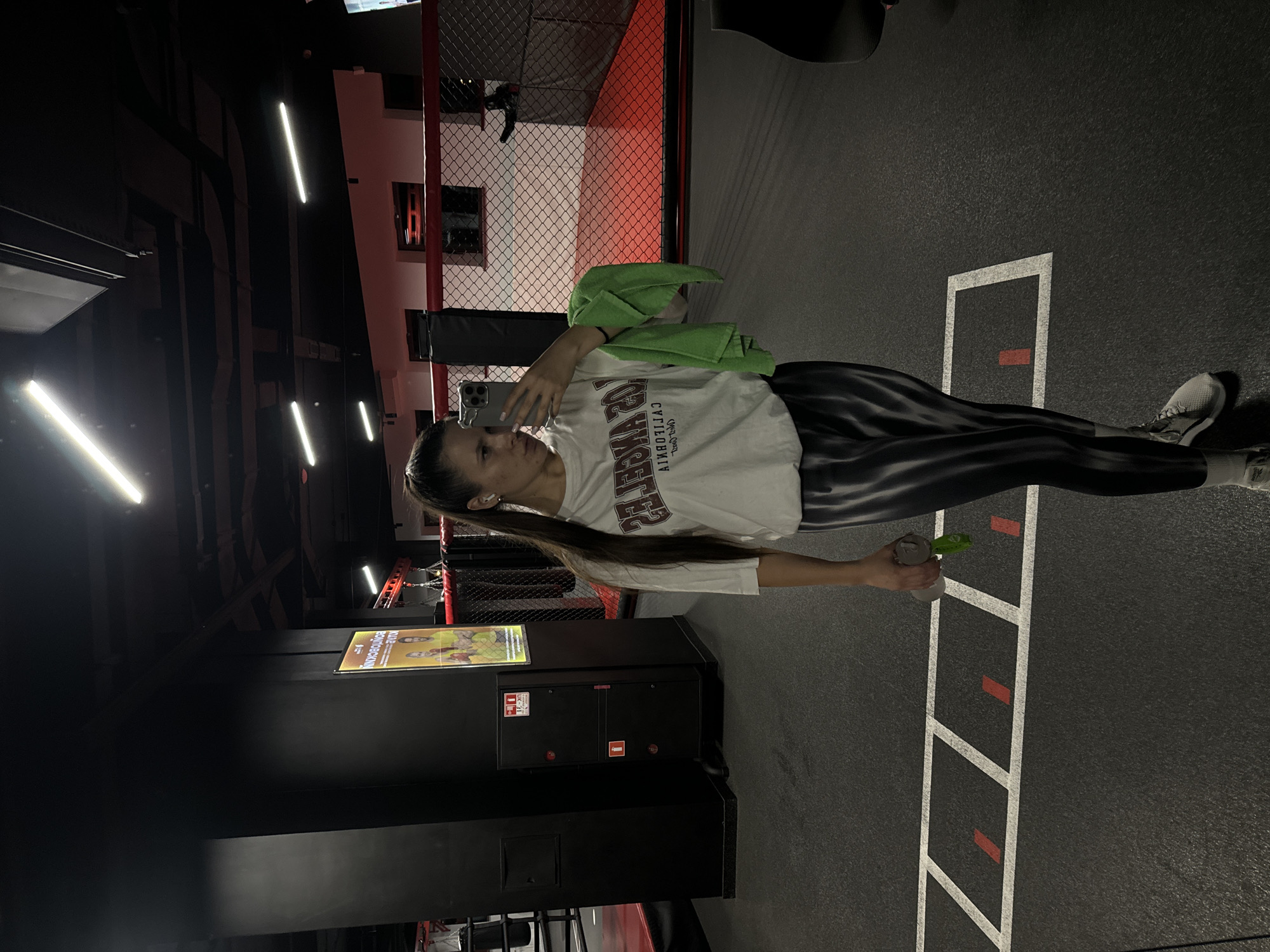 The image shows a woman in what appears to be a gym or fitness center located in Vniissok, Russia. The floor is black with white lane markings. She is wearing a white t-shirt, striped leggings, and sneakers. A green towel is draped over her shoulder, and she holds a spray bottle. She's holding a cell phone, possibly taking a selfie. In the background are gym equipment, black lockers or cabinets, and overhead lighting. A fenced enclosure suggests a training area. A poster is visible on one of the cabinets. The area appears dimly lit.