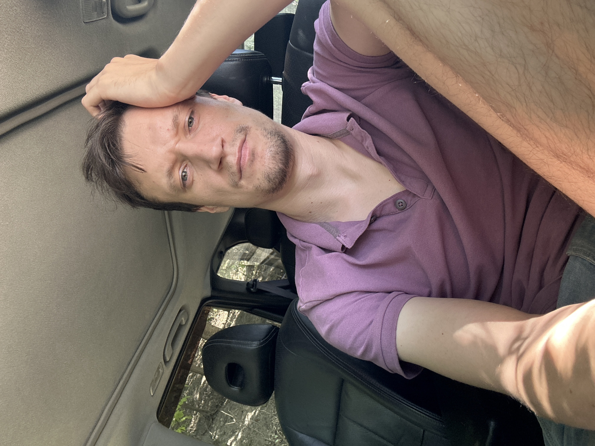 A man is seated in the driver's seat of a car, with his right hand resting on his forehead. He is wearing a purple polo shirt. The car's interior is visible, including black leather seats and a sunroof. Outside the car, trees and foliage can be seen through the windows. The image appears to have been taken during the daytime, with sunlight casting shadows.