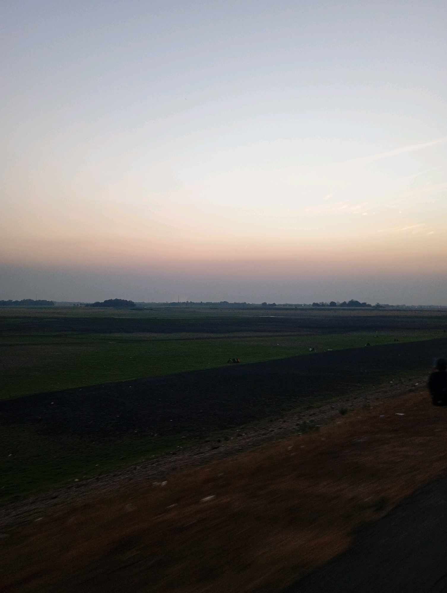 The image depicts a landscape at dusk in Haurkolot, Indonesia. A vast expanse of fields dominates the scene, with some areas appearing as tilled dark soil and others as green vegetation. In the distance, a treeline and small structures are visible, suggesting a rural or semi-rural setting. The sky above is a gradient of soft colors, transitioning from a pale blue at the top to a pale orange near the horizon, indicating the setting sun. The foreground is blurred due to motion, with a dark embankment running diagonally across the bottom right. A lone figure is partially visible in the far right of the frame, their silhouette suggesting they are seated or standing. The overall impression is one of a peaceful, rural evening in Haurkolot.