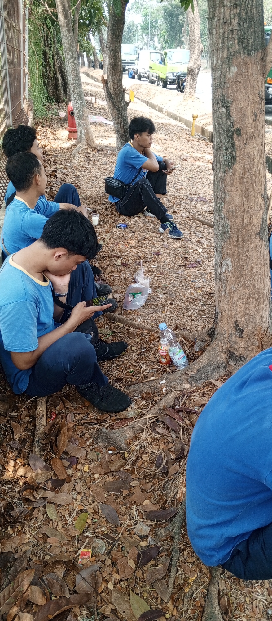 In an outdoor urban setting, a group of young males, likely students given their matching blue shirts, are seen sitting on the ground amongst fallen leaves and tree roots. The foreground is dominated by the legs and lower torsos of two individuals, one of whom is actively engaged with a mobile phone. The midground shows several other young men in similar attire, some looking into the distance, one appearing to be speaking, and another holding a mobile device. Scattered on the ground are various items, including a plastic bag with contents, a bottle of water, a bottle of sauce or drink, cigarette packs, and a small white cup. In the background, trees line a dirt path, and beyond that, several trucks are parked along a road, suggesting a location near a commercial or industrial area. A red fire hydrant is visible to the left, partially obscured by foliage and a fence. The overall impression is a casual gathering during a break or waiting period in a somewhat unkempt, naturalized urban environment.