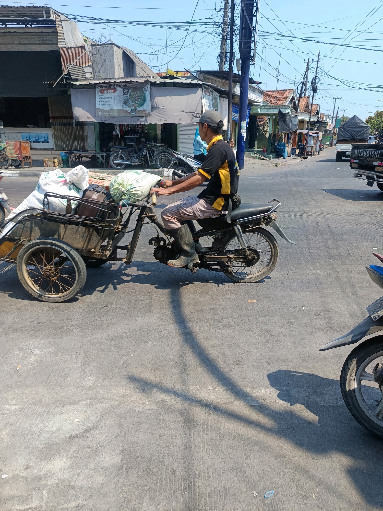 In Blega, Indonesia, a man is riding a motorcycle with a sidecar filled with large white sacks. The man is wearing a grey baseball cap, a yellow and black short-sleeved shirt, and dark boots. The sidecar is attached to the right side of the motorcycle and appears to be carrying the sacks and possibly some other items. The motorcycle and sidecar are on a paved road. In the background, there are buildings, a truck with the word "MITSUBISHI" visible, and numerous power lines overhead. The sky is clear and blue. The overall scene depicts a street in a town or city, likely during the daytime, with commercial activity suggested by the buildings and the delivery vehicle.