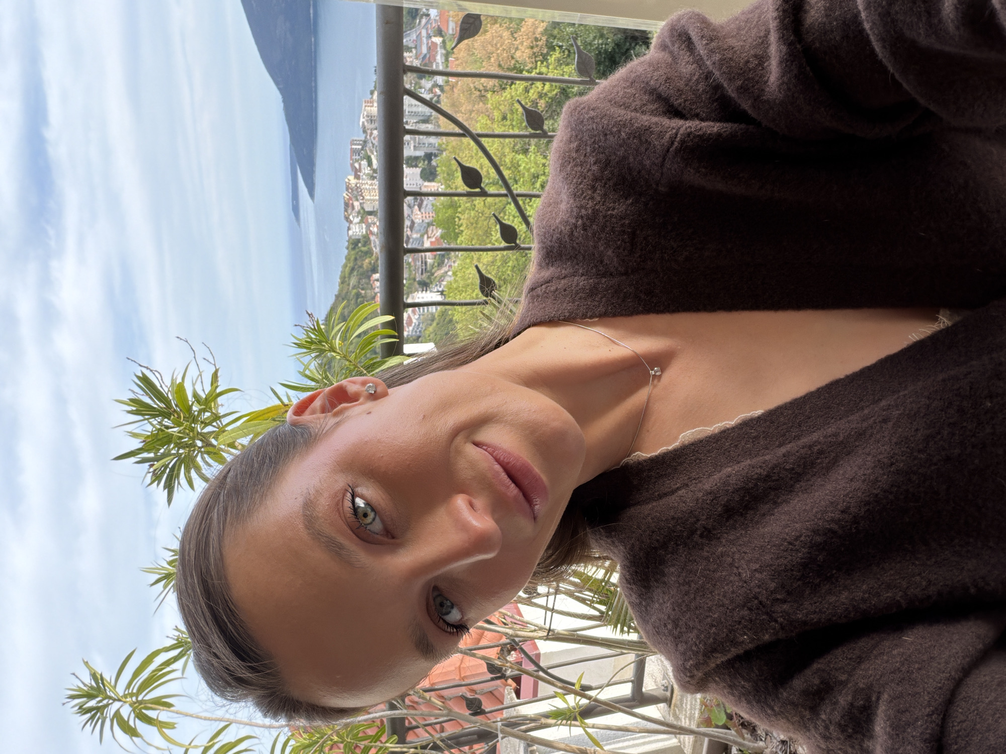 A portrait photograph shows a woman with light skin, brown hair, and green eyes. She is wearing a brown cardigan and a delicate silver necklace. The background reveals a view of the city of Trebesin, Montenegro, with a coastline, buildings, and green vegetation visible through a metal railing with decorative leaf details. A plant with long, green leaves is in the foreground. The sky is partly cloudy.