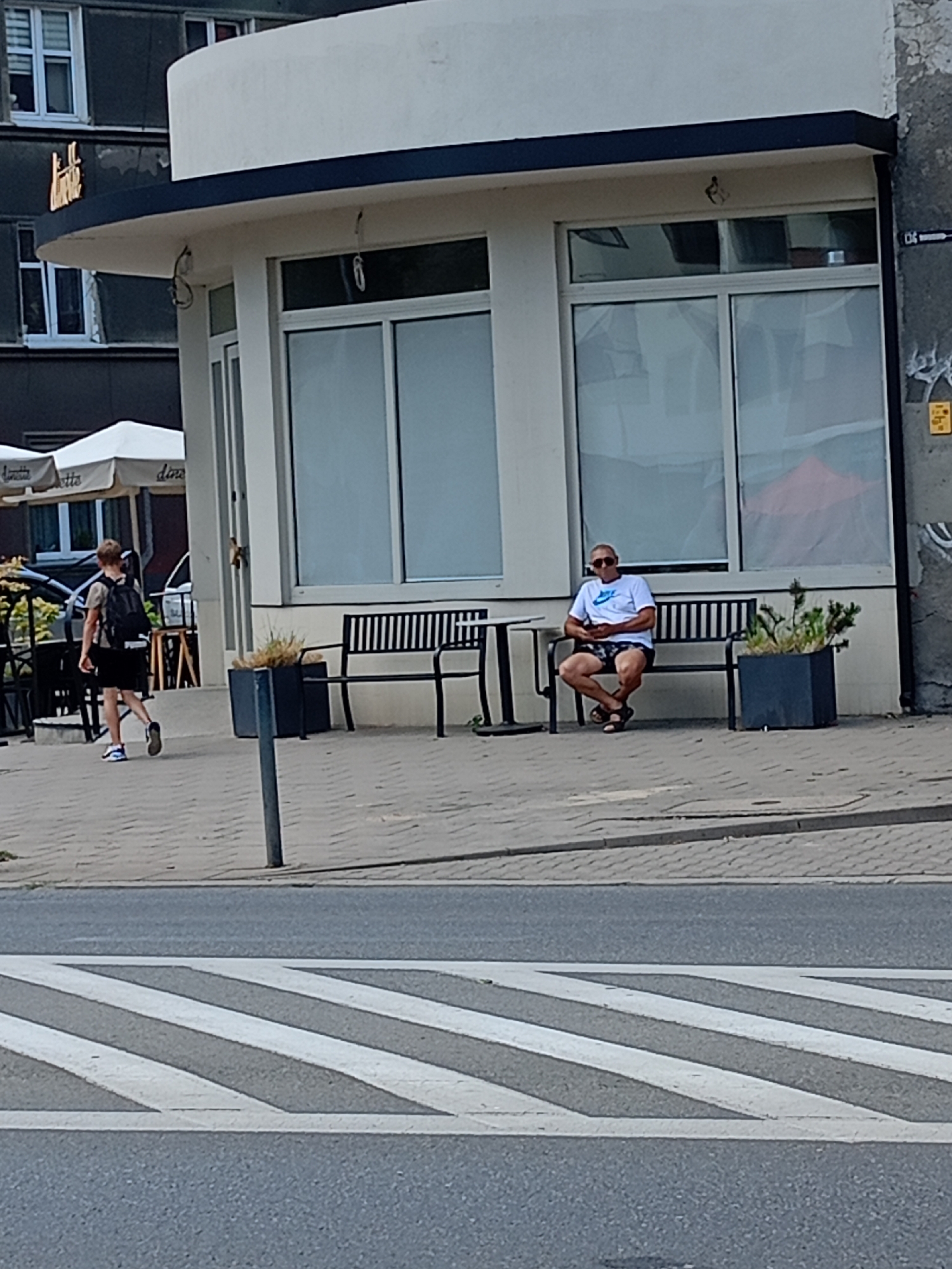In an outdoor urban scene, a young person with blonde hair walks away from the camera towards the left side of the frame. They are wearing a t-shirt, dark shorts, and a backpack. To the right, a man with sunglasses is sitting on a dark bench. He is wearing a white t-shirt and patterned shorts. The bench is in front of a building with large windows. In the foreground, a pedestrian crossing with white stripes is visible on the asphalt. The building has a white curved facade and a dark trim. There are also outdoor umbrellas and tables to the left of the building. The overall environment appears to be a city street or sidewalk area.