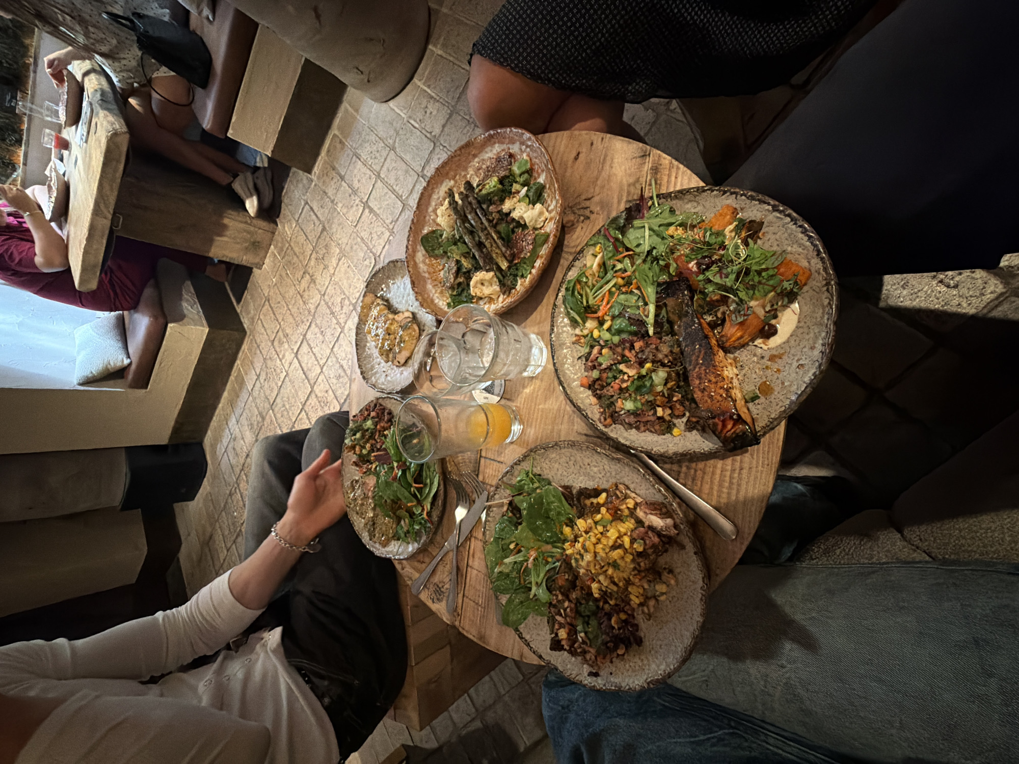 The scene captures a top-down view of a wooden circular table set with several plates of food, likely in a restaurant or dining establishment. Four people are seated around the table, partially visible in the frame.
On the table, there are at least four distinct plates, each containing different food items. One plate appears to have a salad-like arrangement with green leaves, possibly spinach or arugula, and other vegetables, with a reddish-brown ingredient. Another plate contains what looks like grilled asparagus and some white, possibly dairy-based, elements. A third plate has a mixture of grains or rice, and what seems to be corn or another yellowish vegetable. The fourth plate is slightly smaller and contains a golden-brown sauce, possibly on small food pieces.
There are three glasses, two of which appear to be filled with water, and one contains a bright orange liquid, possibly juice. There are also eating utensils, including forks and possibly a knife, placed near one of the plates.
The environment shows some variation. The floor appears to be tiled, with a light beige or tan color. Surrounding the table are seating arrangements. On one side, there's what looks like a bench with light-colored padding. On another side, someone is wearing jeans. On the far left of the image, there's a dining area on a raised platform with stone-like furniture.
The image doesn't explicitly state that the location is in London, United Kingdom, but given the dining setting and the fact that you included that information, one can presume that the place is a restaurant.