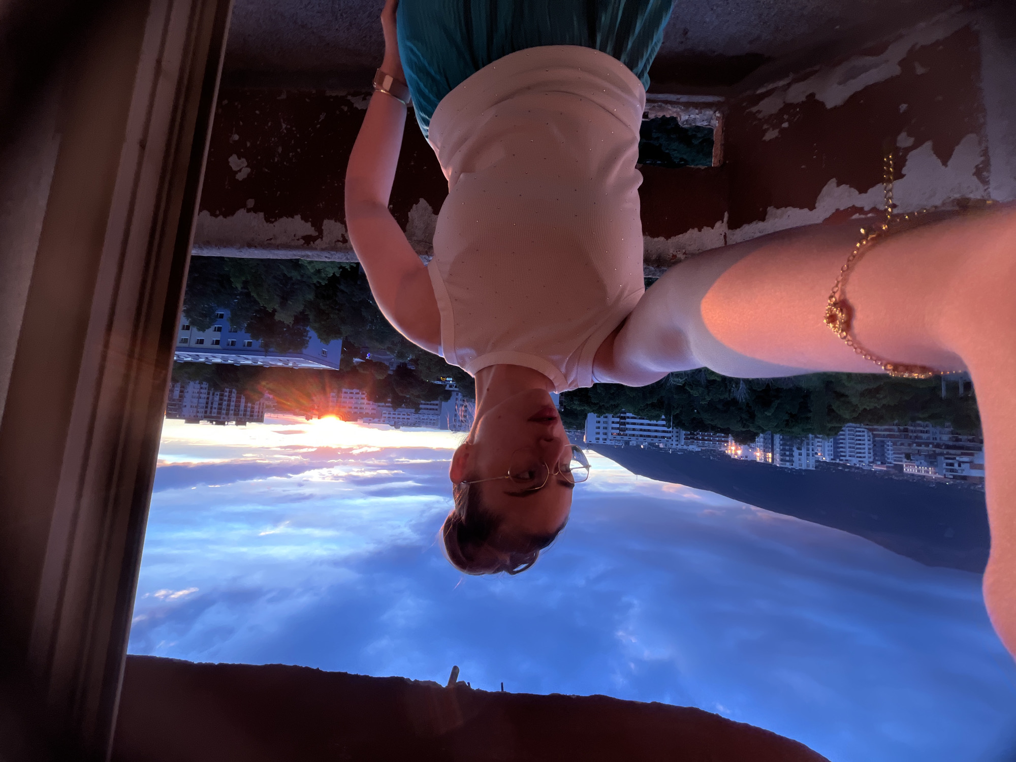 A young woman with her hair in a bun and wearing glasses takes a selfie with her left arm extended. She is wearing a white, sleeveless top with subtle embellishments and a turquoise skirt. A gold bracelet adorns her left wrist. She is standing on a balcony with a weathered railing, overlooking a city at sunset. The sky is a deep blue with streaks of orange and pink from the setting sun, which is visible behind buildings and trees. A mountain range is silhouetted in the distance. The scene is lit by the warm glow of the sunset.