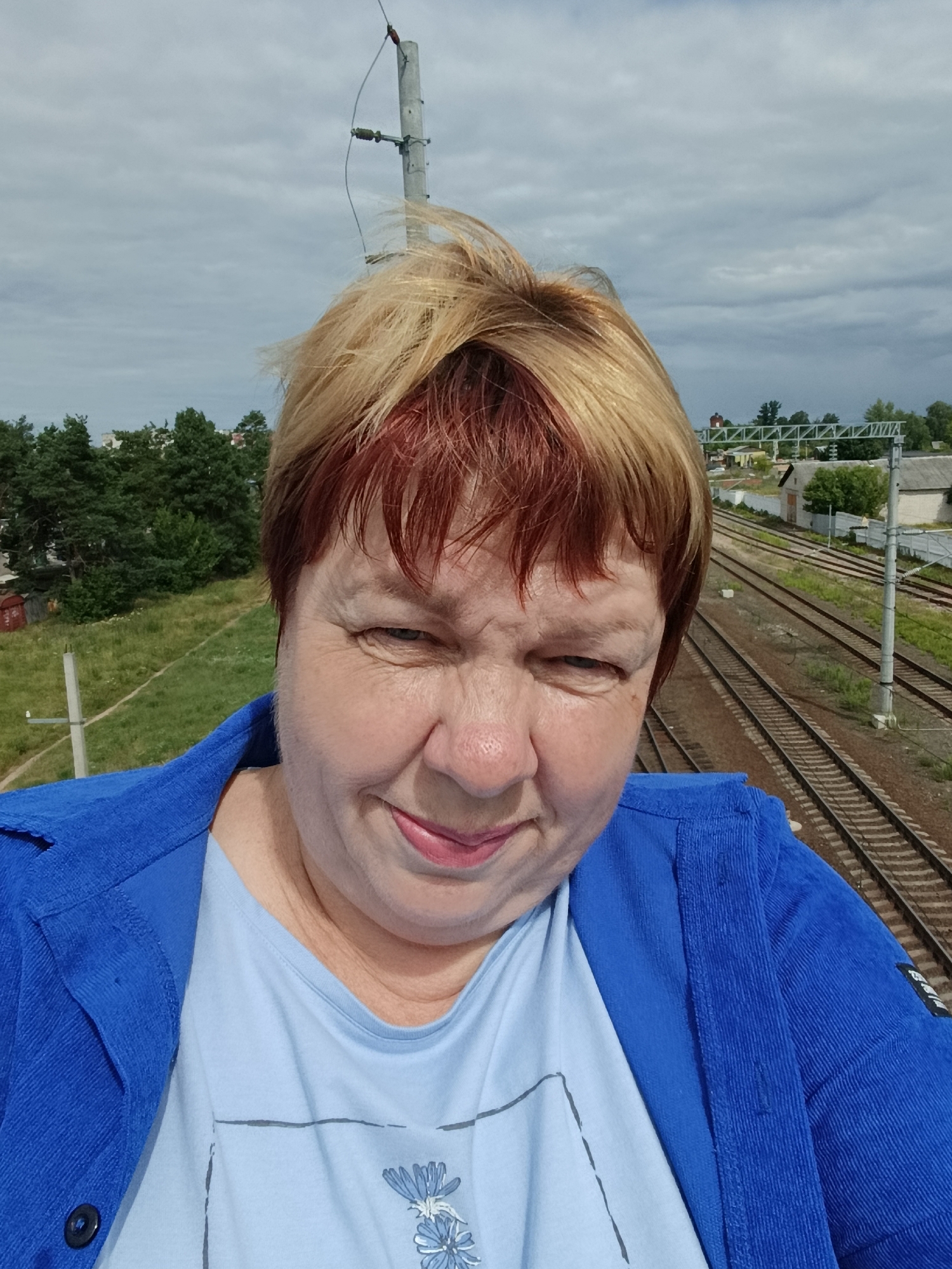 An outdoor, eye-level, medium selfie captures a woman with short, blonde and red-tinted hair. She is wearing a blue collared jacket over a light blue t-shirt. Her expression is a slight smile with her eyes squinted. The background reveals a cloudy sky and a wide view of multiple train tracks, suggesting a location near a railway. The environment appears to be a town, with some trees and buildings visible in the distance, consistent with the city of Svietlahorsk, Belarus, known for its industrial areas and railway infrastructure. A utility pole stands prominently in the frame behind the woman's head.