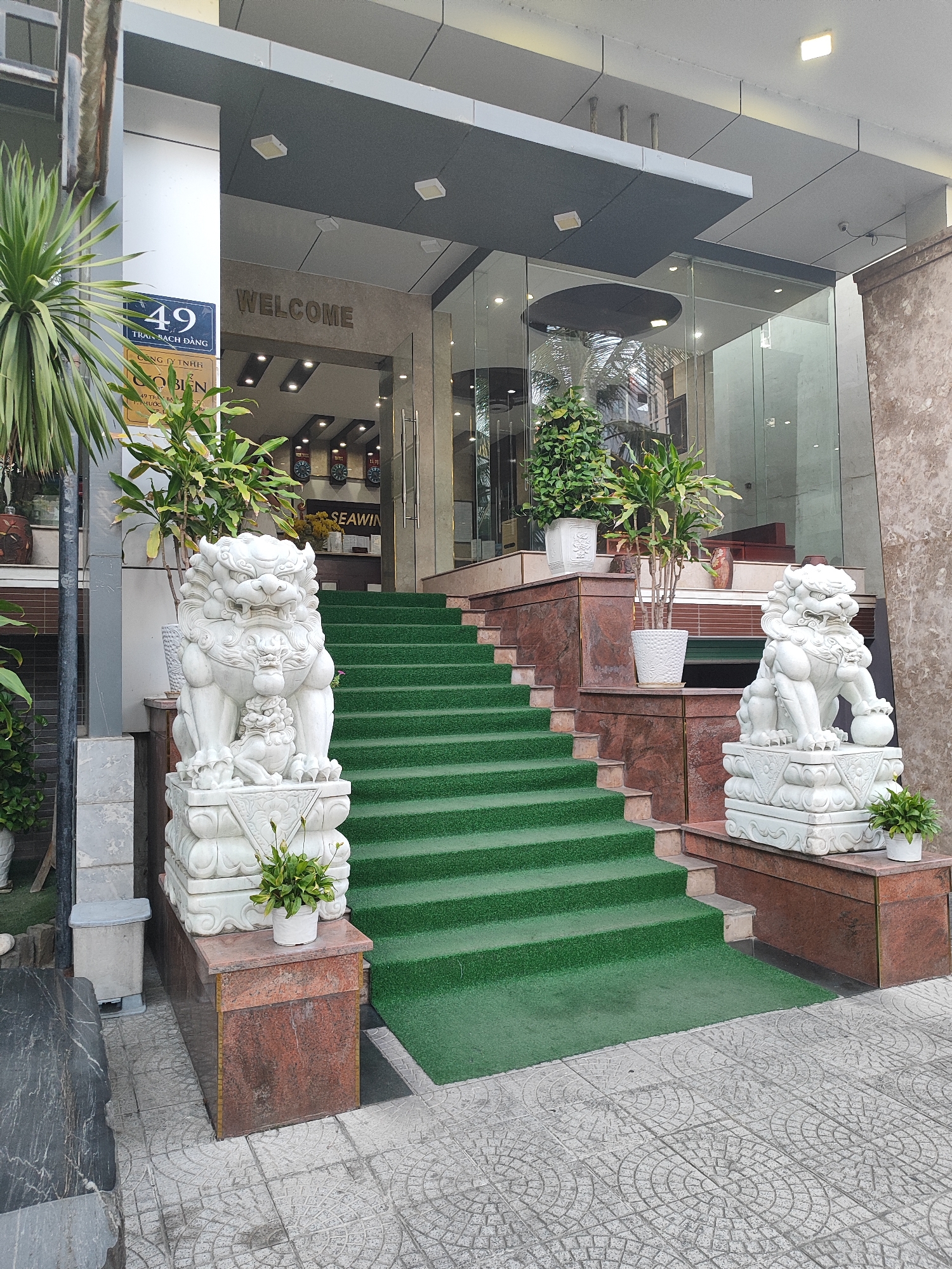 This media file depicts the entrance of a building in Đà Nẵng, Vietnam. The scene features a prominent set of green carpeted stairs leading up to a glass-enclosed entrance. Flanking the base of the stairs are two white stone statues of Foo dogs, traditional Chinese guardians. The left statue is positioned on a reddish-brown granite base, next to a potted plant and a sign that reads "49 TRẦN BẠCH ĐẰNG" and "CÔNG TY TNHH NEOBLEN". The right statue sits on a similar base, also with a potted plant.
Above the entrance, the word "WELCOME" is inscribed in a muted tone. The building's facade is modern, with large glass panels reflecting the surroundings. Inside, a reception area is visible, with a counter and what appear to be clocks on the wall. The lighting suggests it is daytime, and the weather is clear. The overall impression is one of a well-maintained, possibly commercial or hotel establishment, with an emphasis on traditional decorative elements at its entrance.