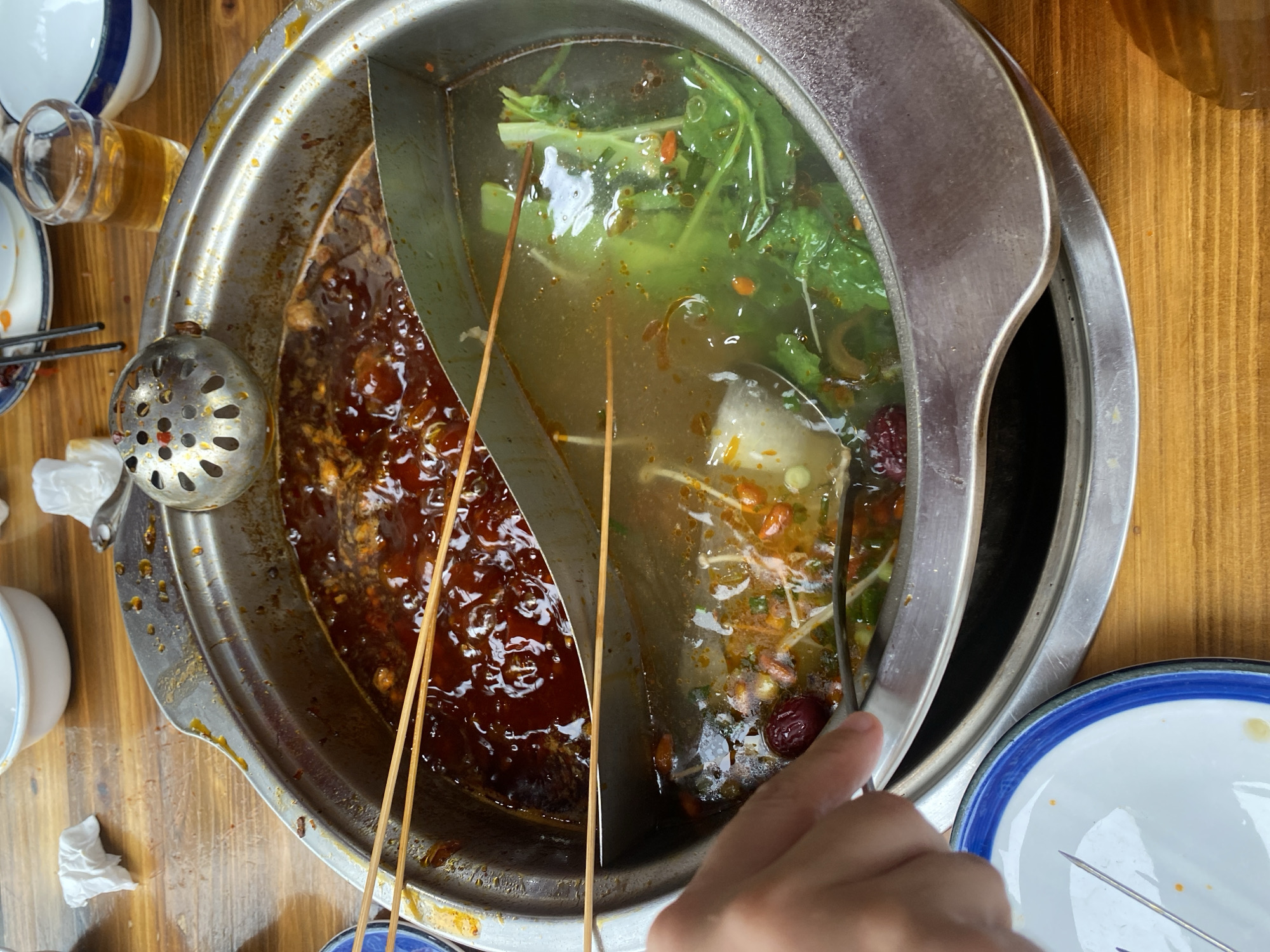 The image shows a close-up, overhead view of a split hot pot on a wooden table. The hot pot is divided into two sections: one filled with a dark red, oily broth with visible pieces of meat and spices, and the other with a lighter broth containing green vegetables, mushrooms, and what appear to be dates. Several wooden skewers are placed across the top of the hot pot, partially submerged in both broths. A metal ladle with perforations rests on the edge of the hot pot. A human hand is visible on the left side of the image, holding a pair of metal tongs, reaching towards the hot pot. In the background, out of focus, are white ceramic bowls and a small glass of amber liquid. The overall setting suggests a meal is in progress. The image's metadata indicates it was taken in Chongzhou City, China.