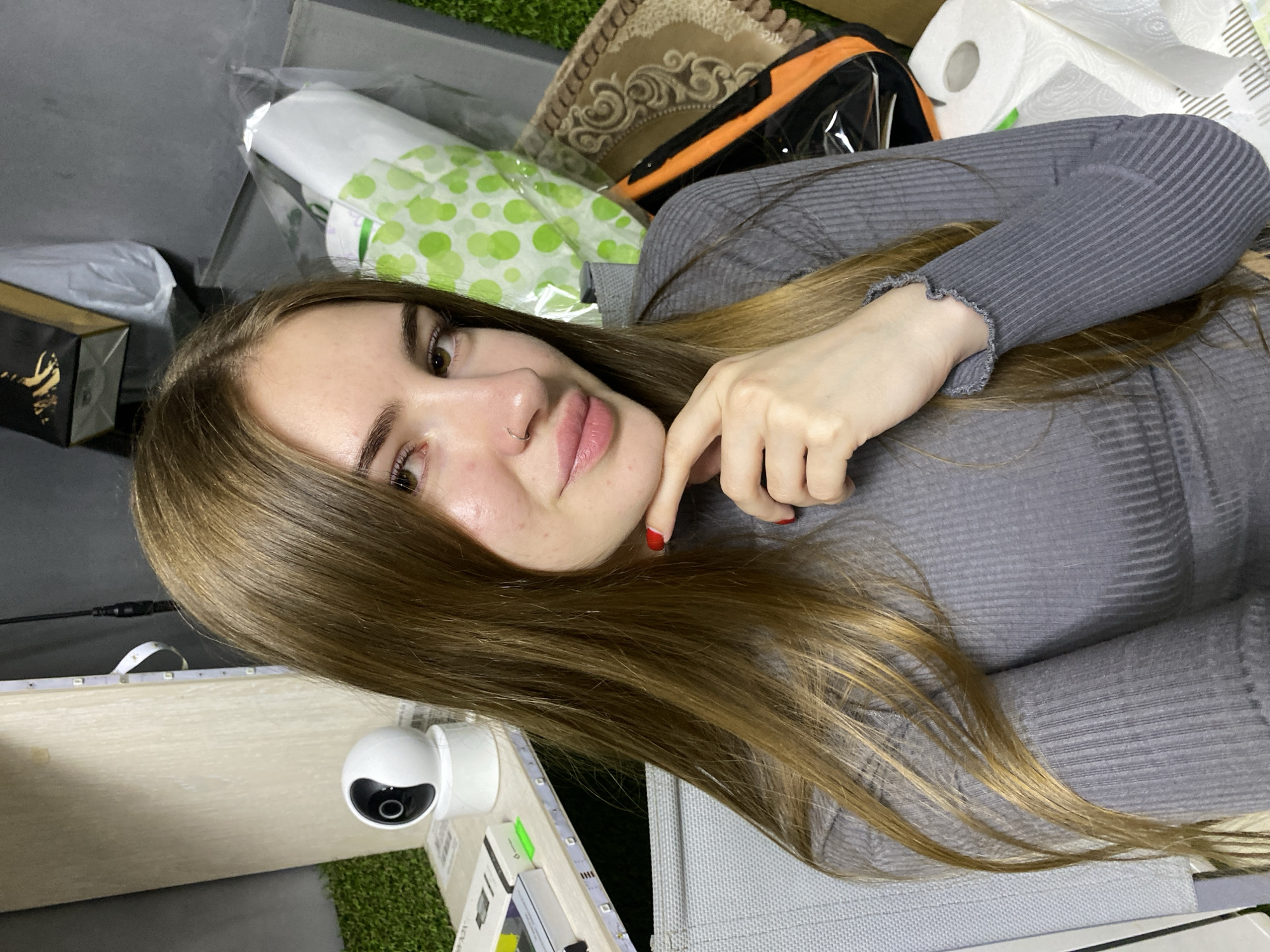 A young woman with long, brown hair is looking at the camera, resting her chin on her hand. She is wearing a grey, ribbed, long-sleeved shirt. She has a nose ring and her lips are slightly parted. The background is cluttered with various objects, including a white security camera on a shelf, packages wrapped in green polka-dot paper, a patterned rug, and rolls of toilet paper. The scene appears to be indoors, possibly a room or storage area. There is no indication of the location being specifically in Dyakovo, Russia.