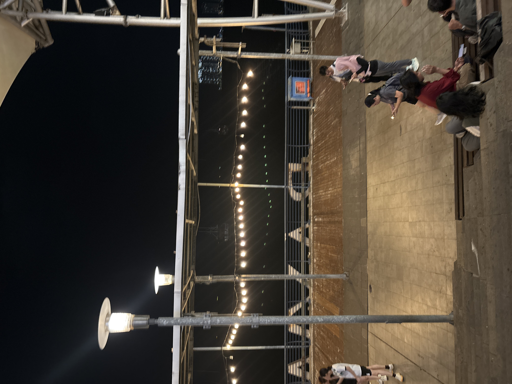 This image depicts an outdoor evening scene on a pier or boardwalk in North Jakarta, Indonesia. In the foreground, several people are visible sitting and standing on tiered steps and the paved walkway. One person in a red top and tan pants is sitting on a bench, holding a phone and appearing to gesture with their hands. To their right, another person in a dark shirt is seated. Behind them, a woman in a grey shirt and black shorts, with a backpack, is looking at her phone. Further back, a person in a pink hijab and grey pants stands while looking at a phone, and another person in a black shirt stands beside them. On the left side of the image, two individuals are standing, one in a white t-shirt and shorts, and the other partially obscured. The boardwalk is illuminated by strings of round lights strung overhead, as well as tall lampposts. Beyond the railing of the boardwalk, the dark water of the sea is visible, with distant city lights and buildings. A blue and orange cooler or utility box is situated on the boardwalk. The overall atmosphere is casual and recreational.