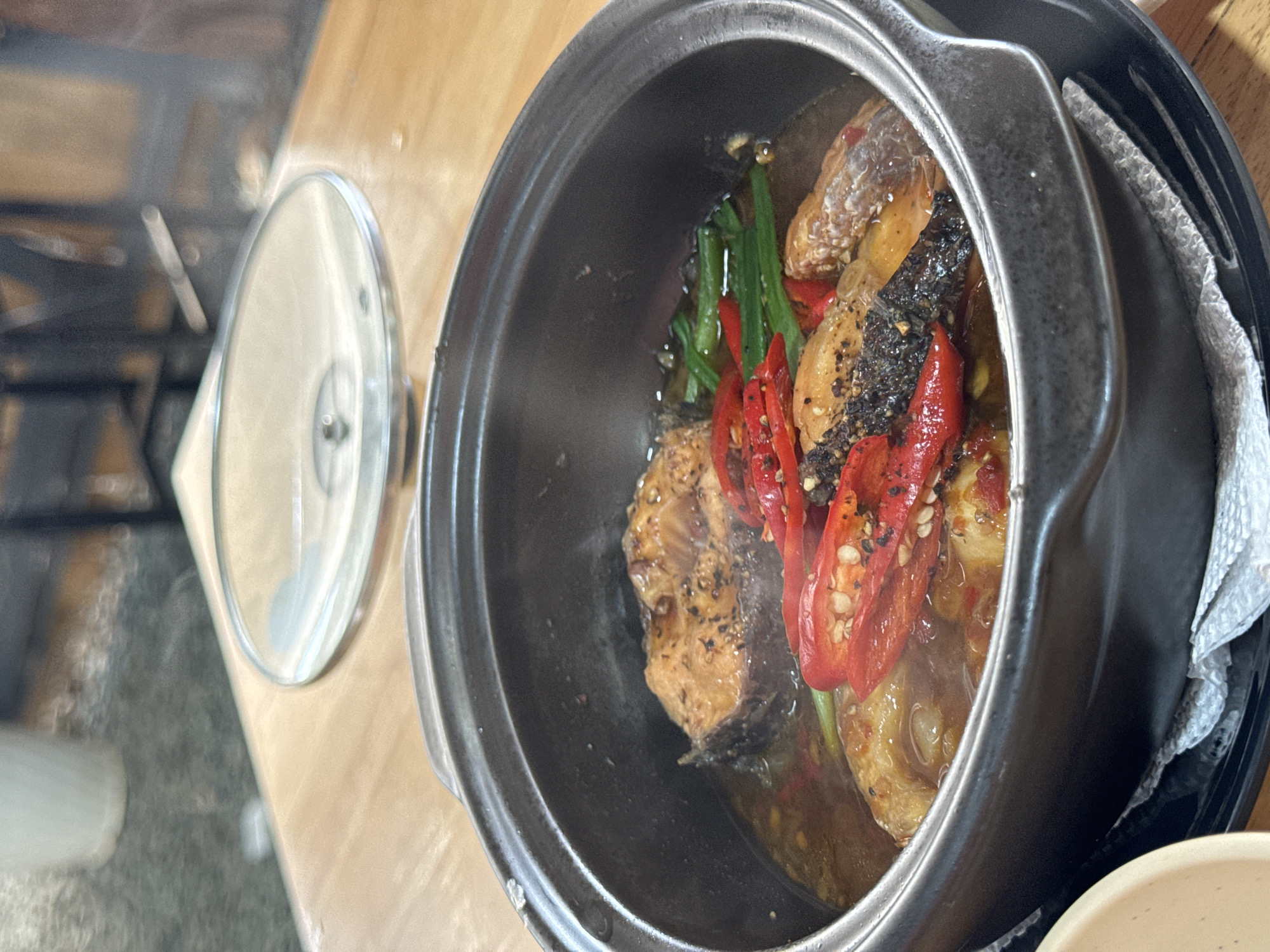 The image captures a culinary scene, likely in a restaurant or home in Thôn 3, Vietnam. A black pot, filled with what appears to be a fish dish, dominates the frame. The dish includes visible pieces of cooked fish, sliced red chili peppers, and green vegetables, possibly scallions. A clear lid rests off to the side, suggesting the dish was recently prepared or is ready to be served. The pot sits on a wooden table. A white cloth or napkin is tucked between the pot and its base, potentially to protect the surface. The scene is well-lit, highlighting the colors and textures of the food.