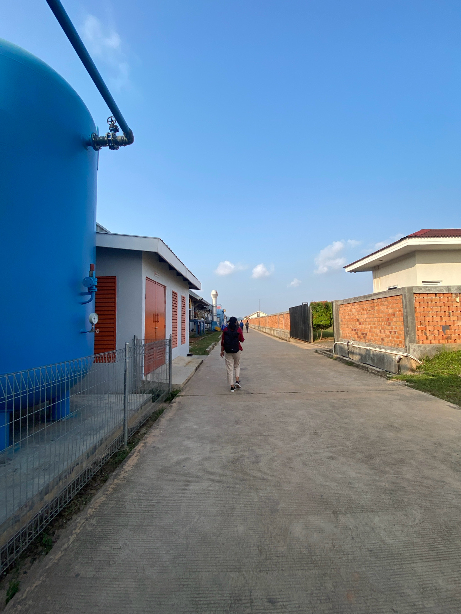 The image shows a person walking away from the camera down a paved path in an industrial area, possibly a water treatment plant or a similar facility. On the left, a large blue tank with pipes and gauges is visible, enclosed by a metal fence. To the right of the path are several buildings with orange doors and windows, a brick wall, and a white building with a red roof. The sky is clear blue with a few scattered clouds. The person is wearing a backpack and casual clothing. The overall environment suggests an outdoor industrial or commercial setting. The exact location in Batam, Indonesia, cannot be definitively determined from the image alone, but the architectural style and industrial infrastructure are consistent with such a location.