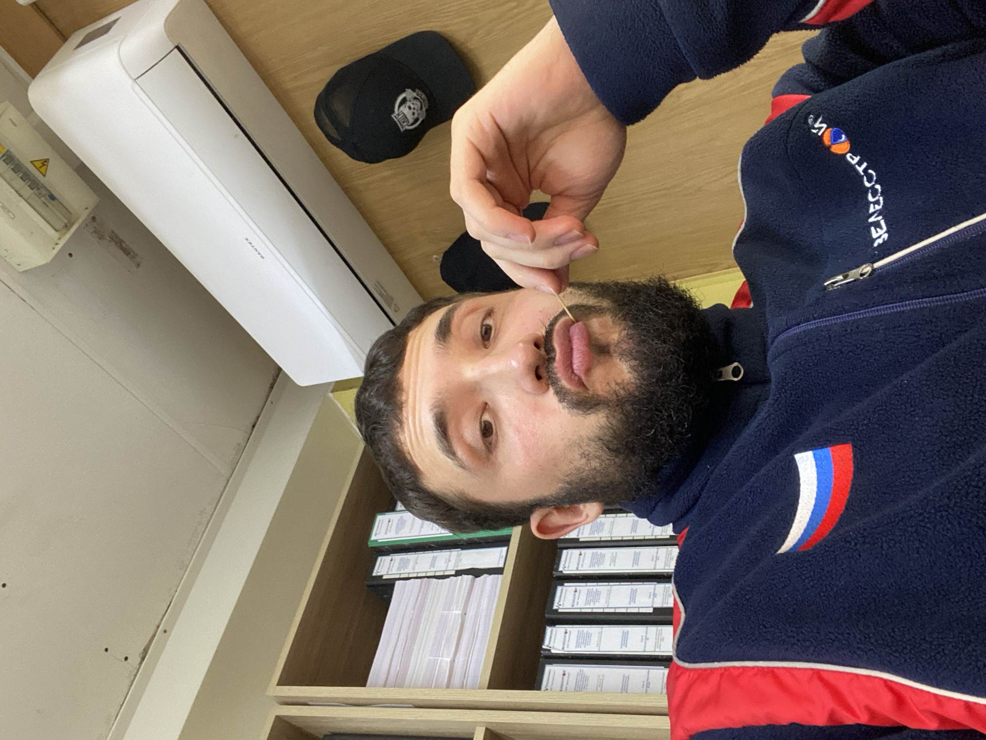 The image shows a man in a dark blue fleece jacket with red and white trim and a Russian flag patch on the left chest. He has a beard and is holding a toothpick to his mouth. In the background, there are shelves with binders, an air conditioner, and a black baseball cap with a skull logo hanging on the wall. The overall setting appears to be an office or storage room. The image does not provide any specific information about the city of Luzhitsy, Russia, or any other geographical location.