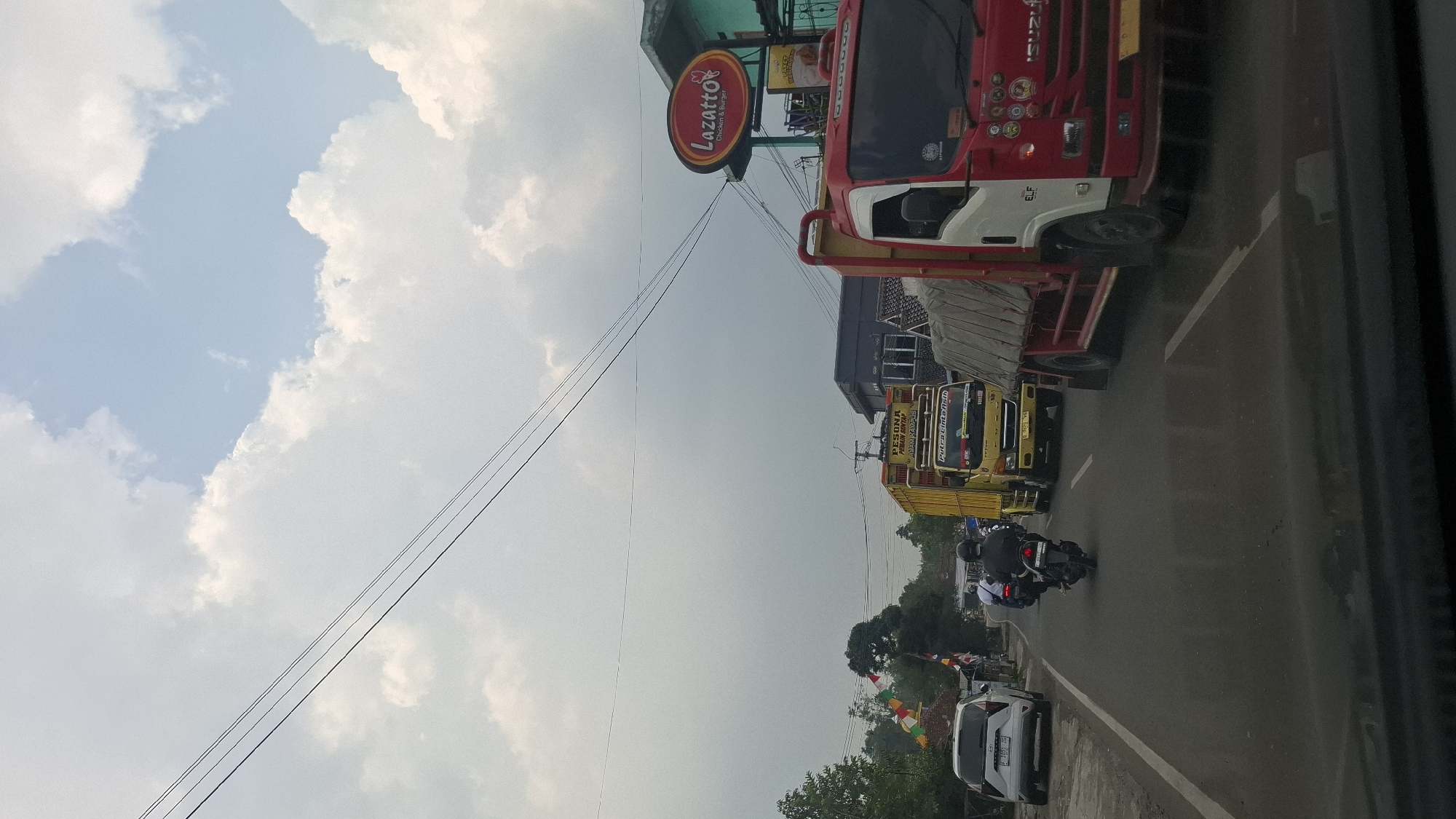 The image captures a traffic scene on a road in Susukan, Indonesia. A white SUV is parked on the left side of the road. A motorcycle with two occupants is in motion on the road, followed by a yellow truck and a red truck. Overhead power lines stretch across the frame. In the background, there are buildings, trees, and a sign that reads "Lazatto Chicken & Burger". The sky is partly cloudy.