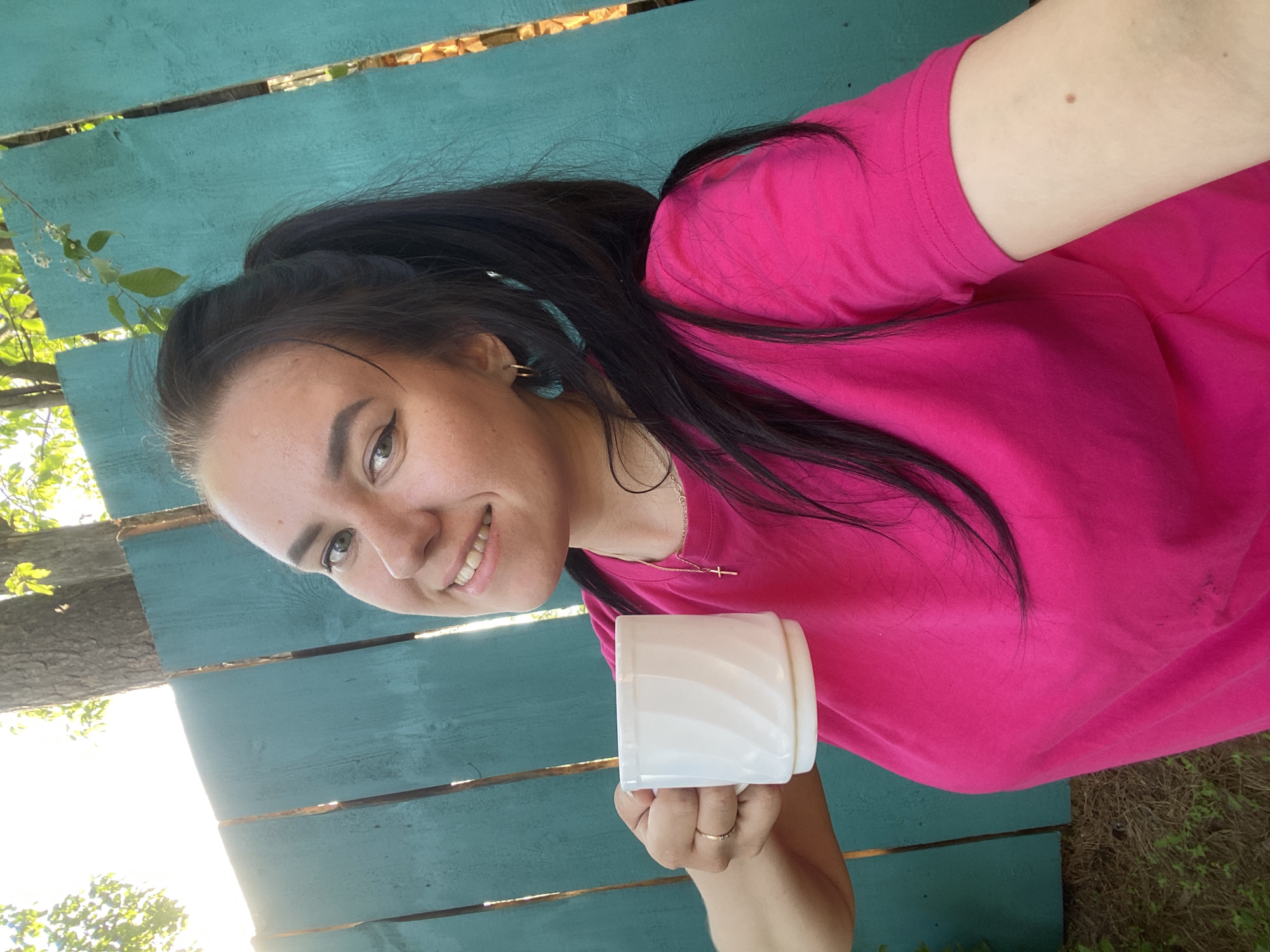 A young woman with dark hair pulled back in a ponytail smiles at the camera while holding a white mug. She is wearing a pink t-shirt and a gold necklace with a cross pendant. She is standing in front of a turquoise wooden fence. The location appears to be outdoors, possibly in a garden or park, with sunlight visible. The city of Chaikovsky, Russia, is not definitively discernible from the image.