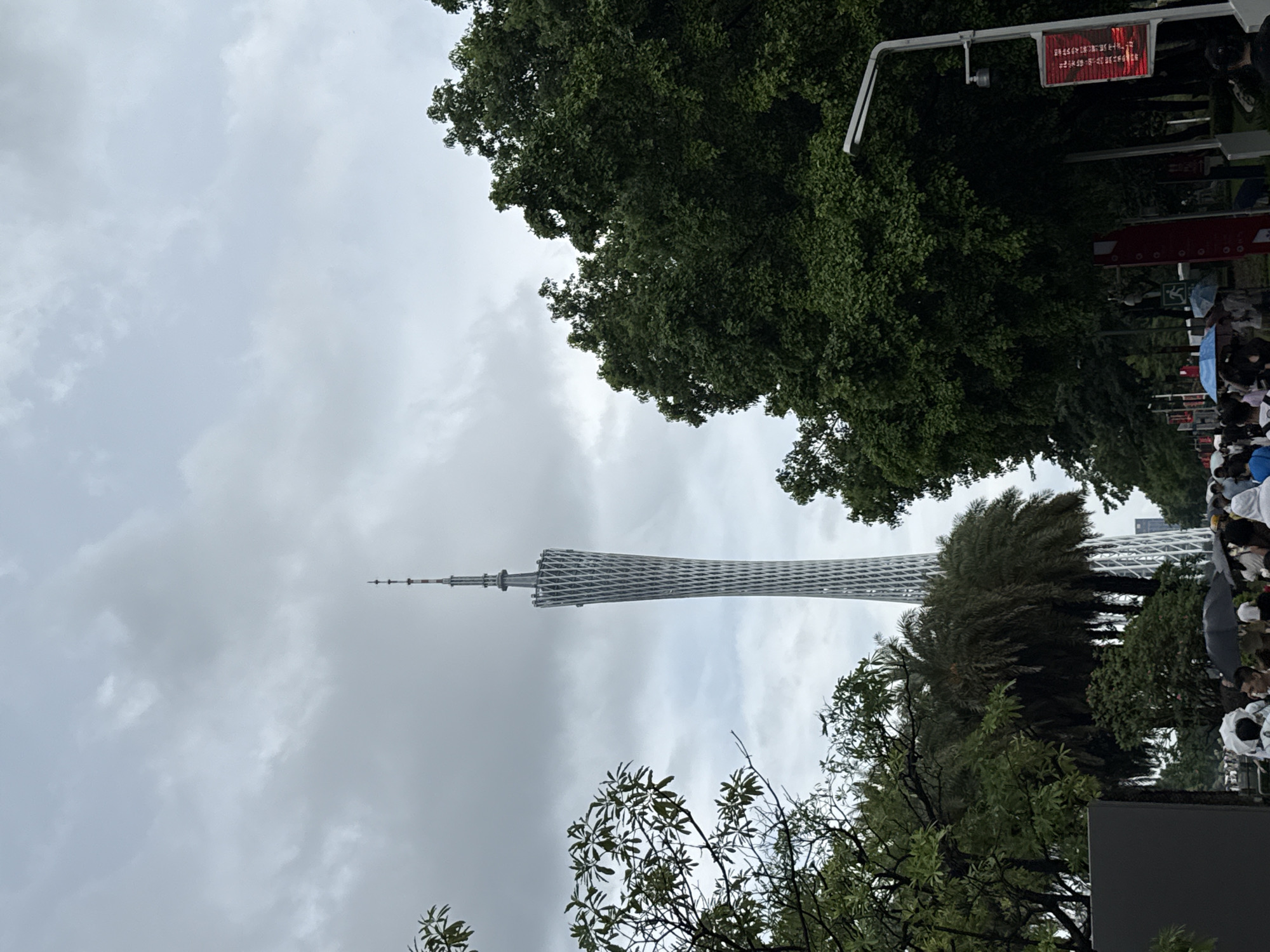 In Tianhe District, China, under a cloudy sky, the Canton Tower, a distinctive lattice-like structure, dominates the background. The tower, made of steel and concrete, tapers towards its apex, which is crowned with antennas. In the foreground, lush green trees frame the scene, with palm trees and deciduous trees intermingling. A crowd of people, some holding umbrellas, are visible, suggesting a potentially wet or overcast day. Streetlights and red banners, possibly displaying advertisements or information, are also present along a pathway. The overall impression is of an urban landscape with a prominent landmark, hinting at a public gathering or a busy thoroughfare.