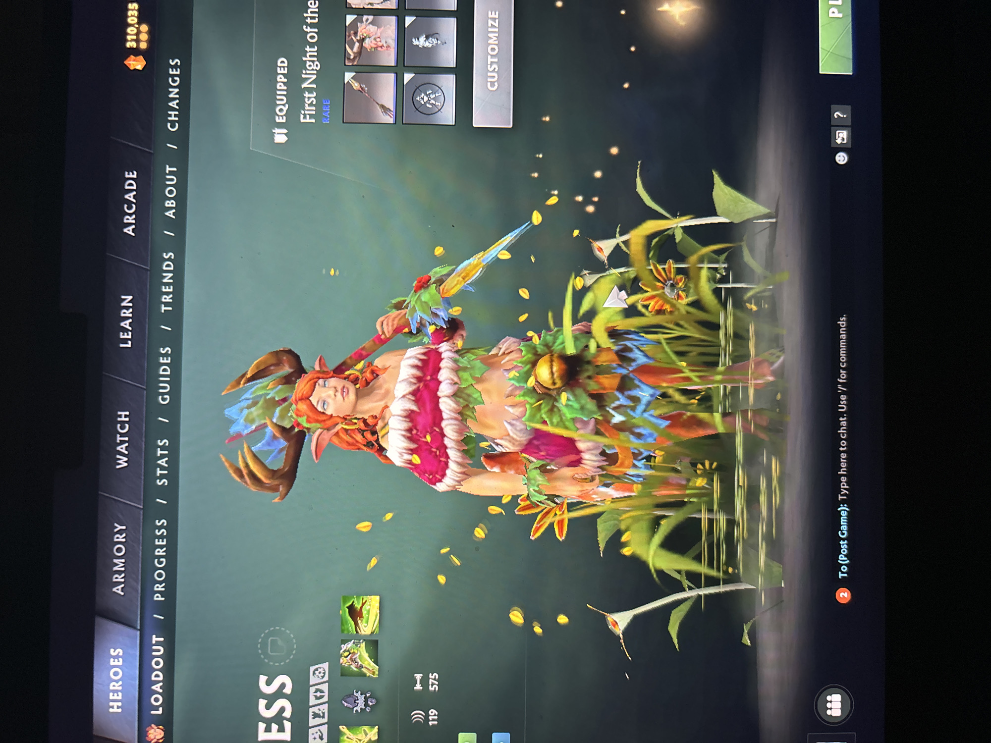 The image displays a vibrant digital fantasy character within a video game's user interface, likely a customization or loadout screen. The central subject is a female character, possibly an elf or nature spirit, featuring reddish-orange hair, pointy ears, and light skin. She wears a costume of lush green leaves, colorful flowers, and vines, including a pink and white leafy top and a green leafy bottom. She holds a glowing, sharp weapon with blue and yellow accents.
The character stands on a dark, reflective surface that mirrors her and the surrounding tall green foliage and yellow-orange flowers. Small glowing, petal-like particles float around her, contributing to an ethereal and magical ambiance.
The game's user interface is visible around the character. On the top left, a currency count of "310,035" is displayed. A vertical navigation menu on the left includes "HEROES," "ARMORY," "WATCH," "LEARN," and "ARCADE." A horizontal sub-menu below reads "LOADOUT / PROGRESS / STATS / GUIDES / TRENDS / ABOUT / CHANGES." Text above the character indicates "EQUIPPED" and "First Night of the," labeled as "RARE," with a "CUSTOMIZE" button nearby. Partial text "ESS" is visible below the character, likely part of her name, along with various game icons and numerical stats ("119," "575"). On the far right, a chat box prompt states "To (Post Game): Type here to chat. Use '/' for commands," accompanied by chat, camera, and question mark icons. A "PLAY" button is partially visible at the top right. This is a digital scene, not a real-world location in Sofia, Bulgaria.