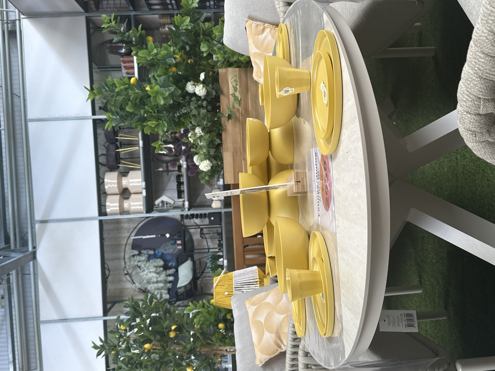 The image depicts an indoor setting, likely a store, displaying outdoor furniture and decor. In the foreground, a round, light-colored table is set with a yellow dinnerware set, including plates and cups. The table is covered with a clear plastic sheet. Behind the table, several yellow bowls are stacked. To the left, a gray armchair with a yellow patterned cushion is visible. Further back, a faux lemon tree with yellow fruit adds a touch of greenery. In the background, shelves are stocked with various items, including decorative vases, planters, and other home goods. The overall environment suggests a retail space, possibly a garden center or home furnishings store, located in Tuk, Netherlands. There are no people visible in the image.