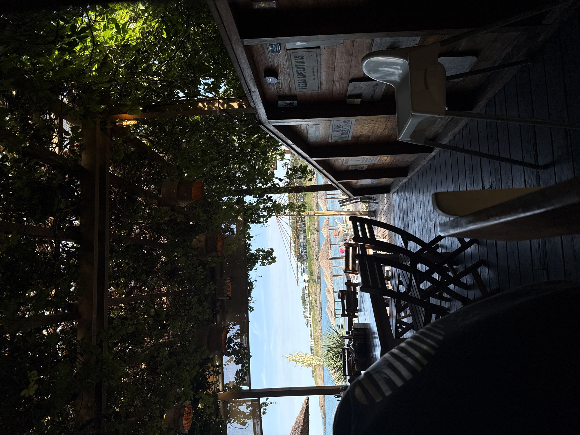 The image depicts an outdoor dining area at a restaurant in Tsarevo, Bulgaria, overlooking the Black Sea. The foreground shows a wooden deck with tables and chairs set for patrons. A portion of a person wearing a dark garment is visible in the lower left corner. To the right, a wooden structure with signs and a high chair is present. In the midground, several thatched umbrellas are arranged on a beach, with the sea and a coastline dotted with buildings visible in the background under a clear blue sky. Lush green foliage drapes over the overhead structure, creating a shaded, natural ambiance.