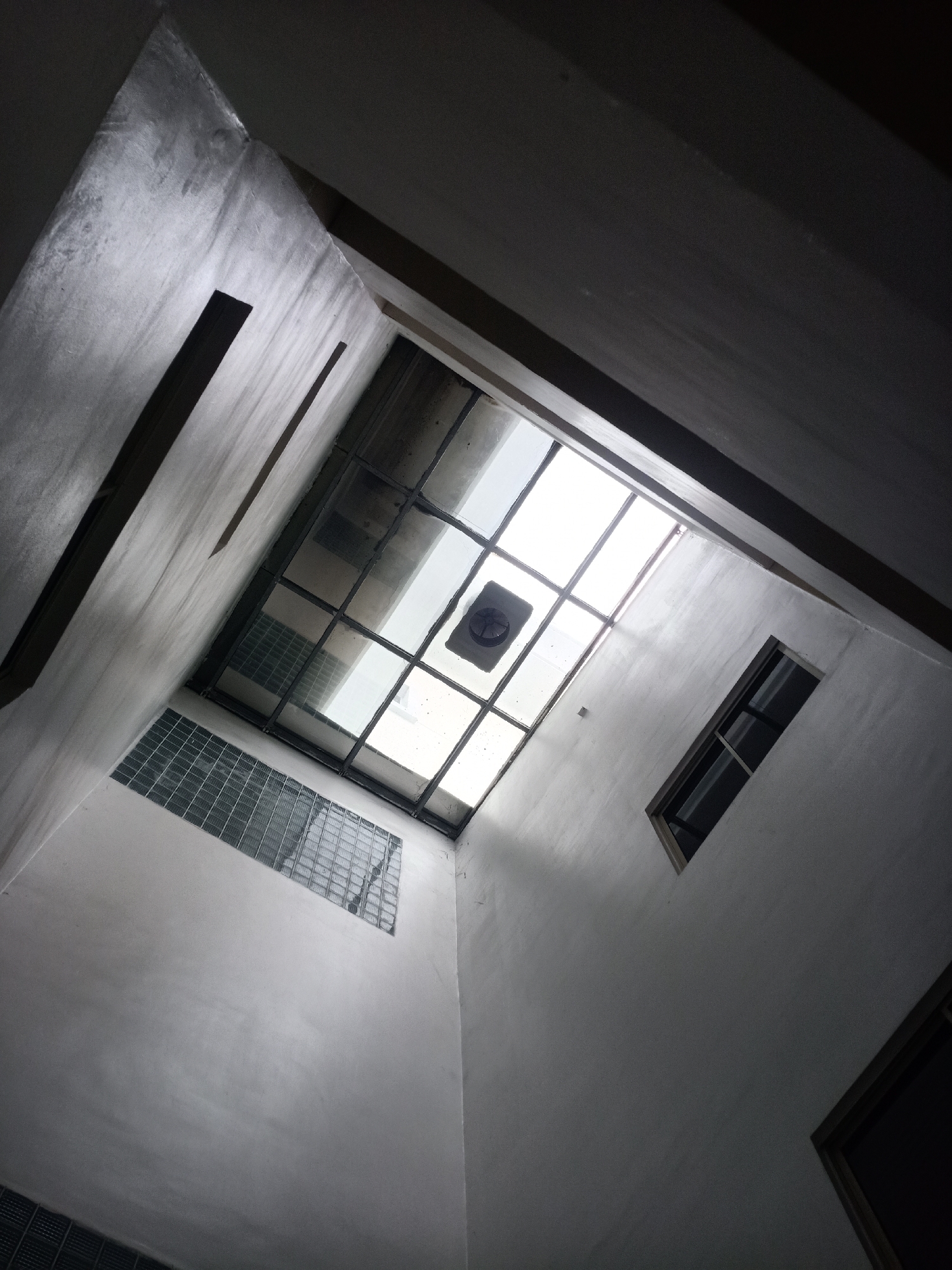 The image depicts a view looking up inside a building, likely a courtyard or atrium. Walls with a rough, textured finish surround a framed glass ceiling. Visible in the ceiling structure is an exhaust fan and daylight coming through. There are also a few windows visible in the walls, positioned at different heights. There are no people or distinct objects to give clues about the specific location or function of the space.